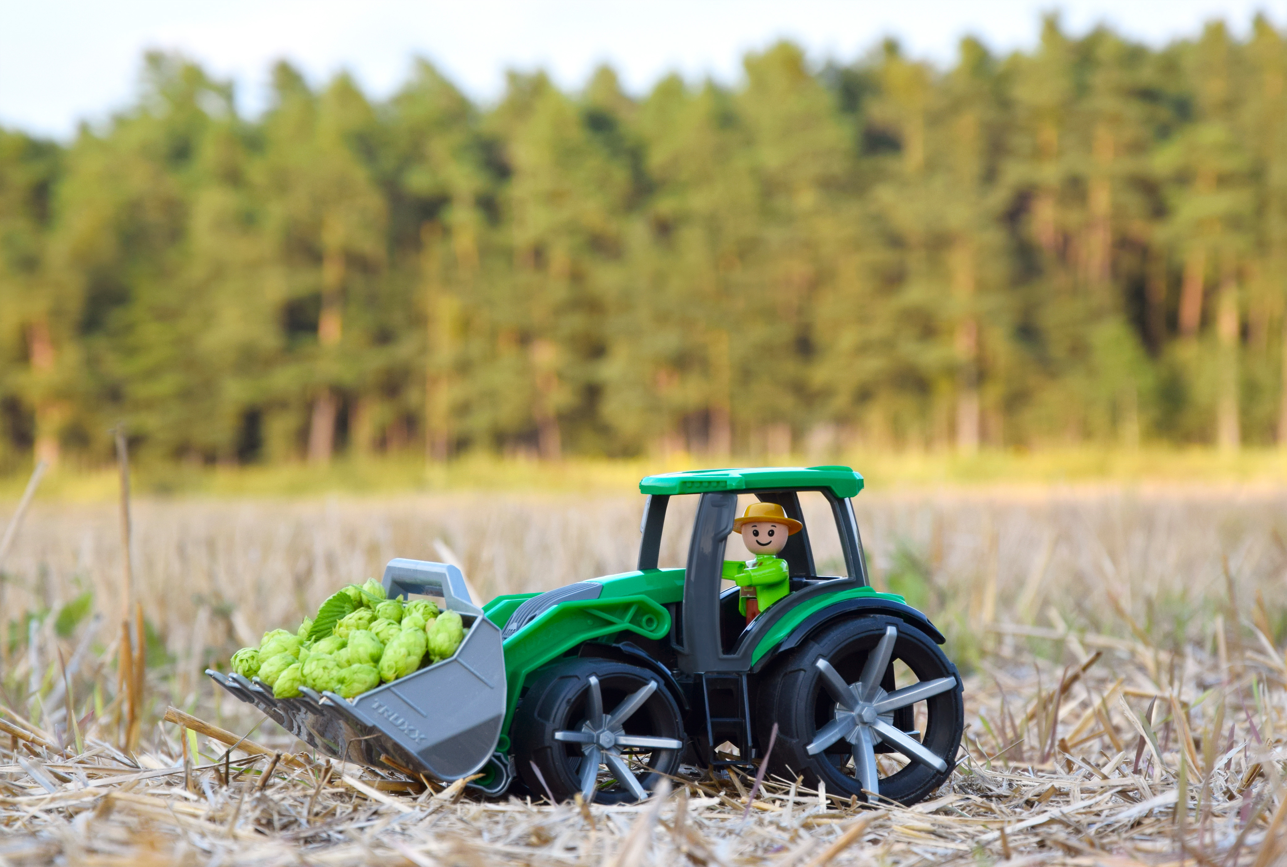 Spielzeugtraktor auf einem Feld, mit einer Figur, einem vollen Eimer und verschwommenem Hintergrund.