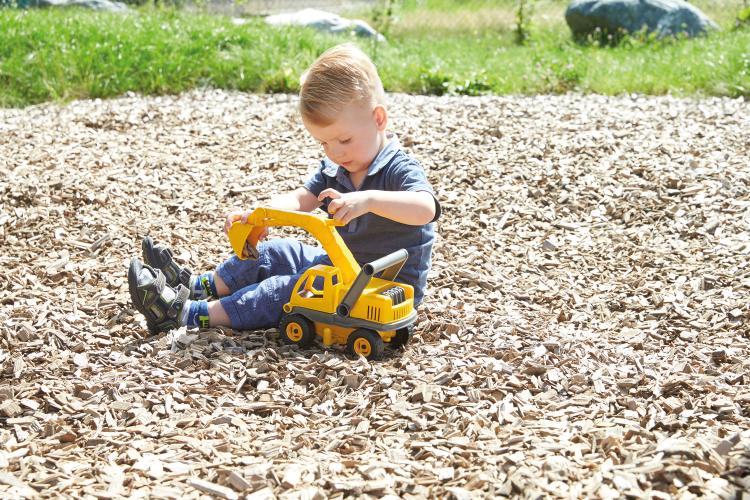 Ein kleiner Junge spielt mit einem gelben Spielzeugbagger auf Holzschnitzeln, grüner Gras-Hintergrund.