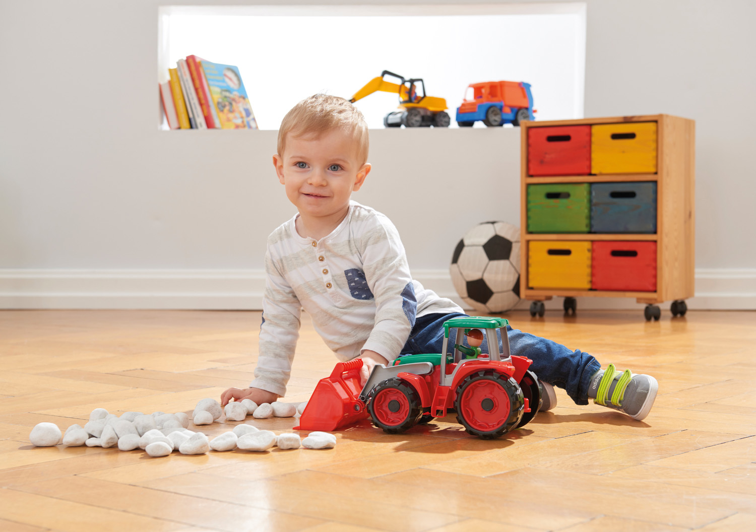 Un niño pequeño juega con un tractor de juguete y piedras. Fondo: juguetes en un estante y una ventana.