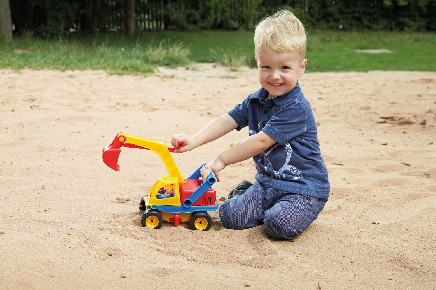 Ein kleiner Junge spielt mit einem Spielzeugbagger im Sand. Hintergrund: Gras und Sand.