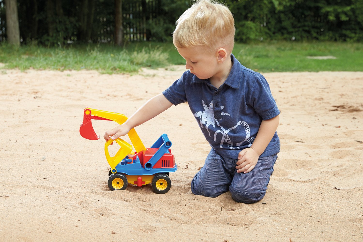Ein Junge kniet im Sand und spielt mit einem Spielzeugbagger. Hintergrund: Sand und Gras.