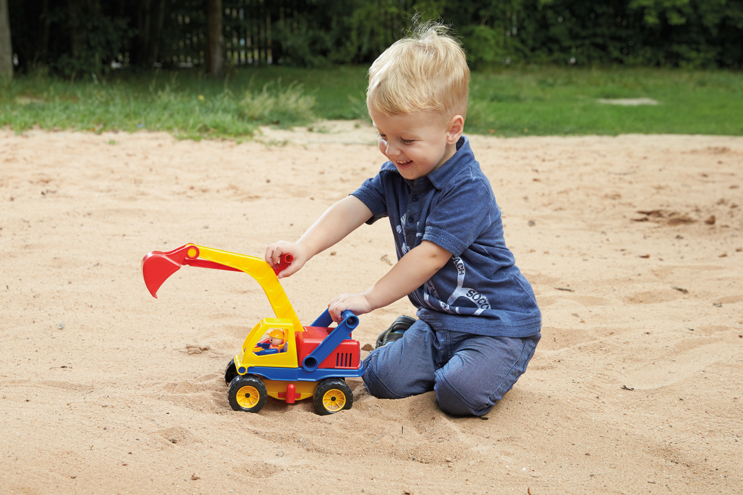 Ein Junge spielt lächelnd mit einem Spielzeugbagger im Sand. Hintergrund: Sand und Gras.