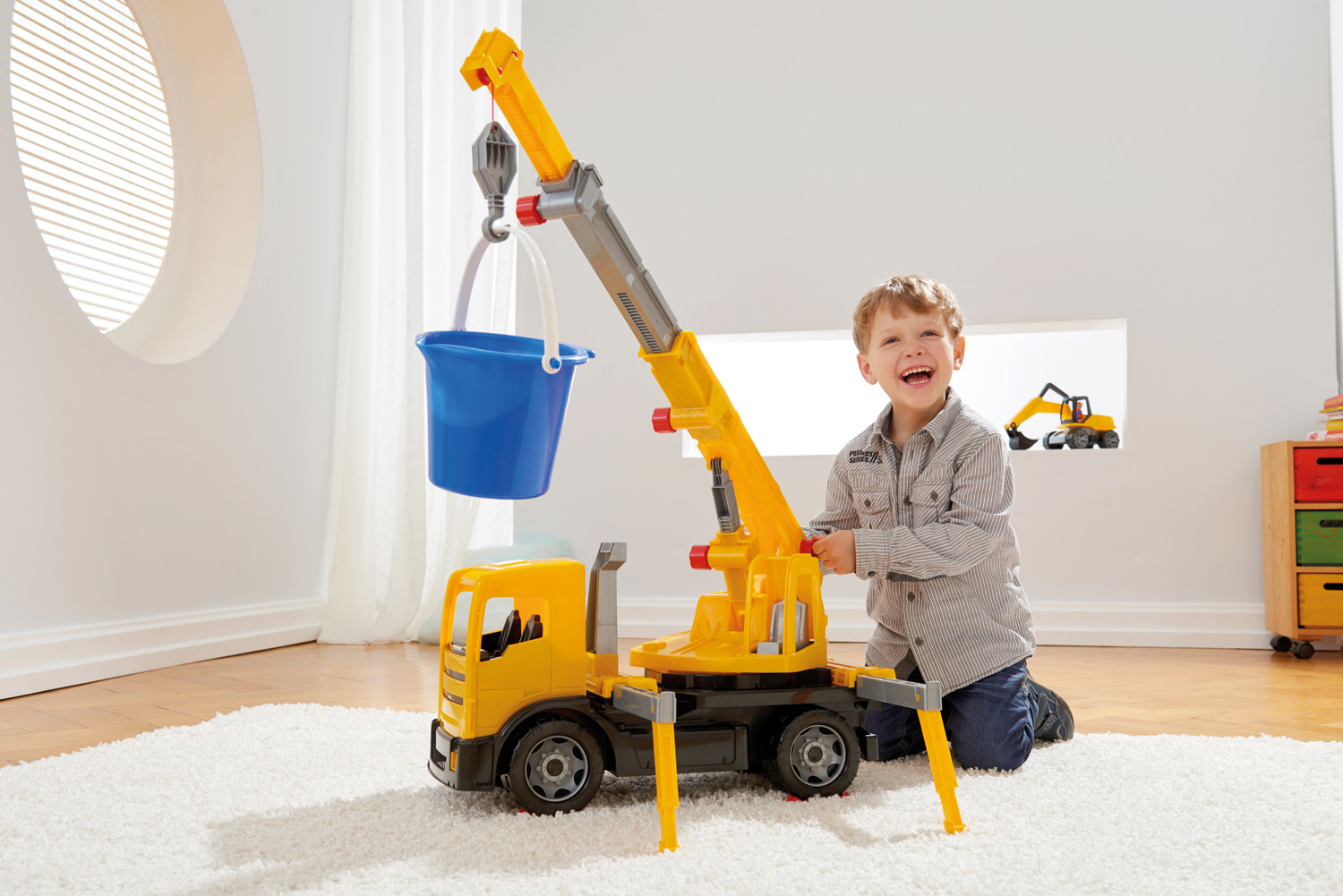 Un bambino sorride accanto a un camion gru giocattolo giallo e grigio. Un secchio blu è appeso alla gru.