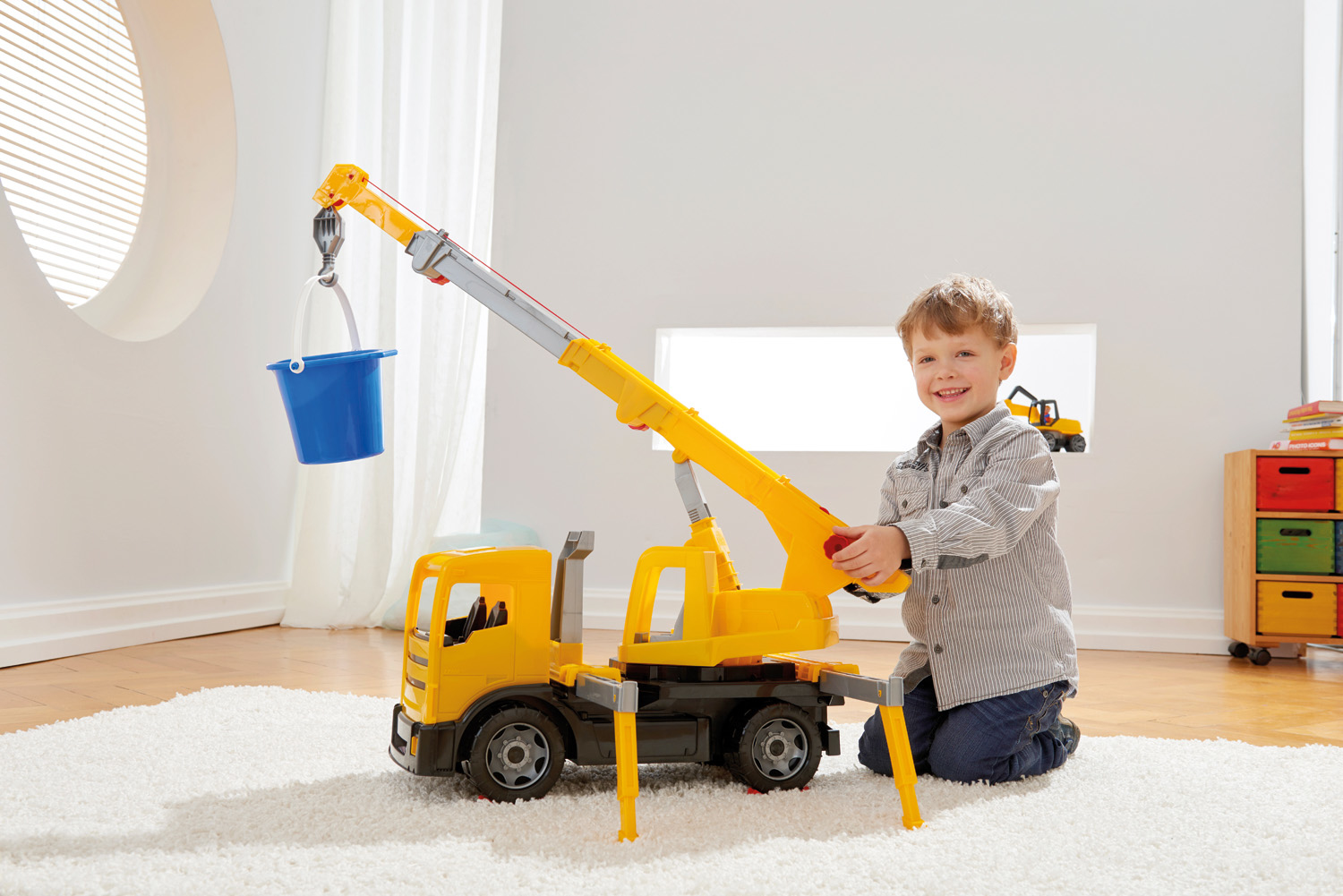 Un bambino sorride, tenendo in mano una gru giocattolo. Un camion gru giallo con un secchio blu è sullo sfondo.