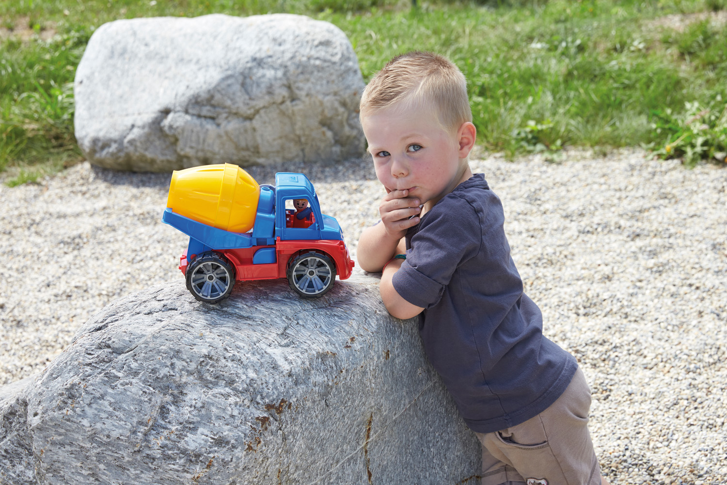 Junge mit blonden Haaren lehnt an einem großen Felsen neben einem Spielzeug-Zementmischer. Grüner Rasen und Kies im Hintergrund.