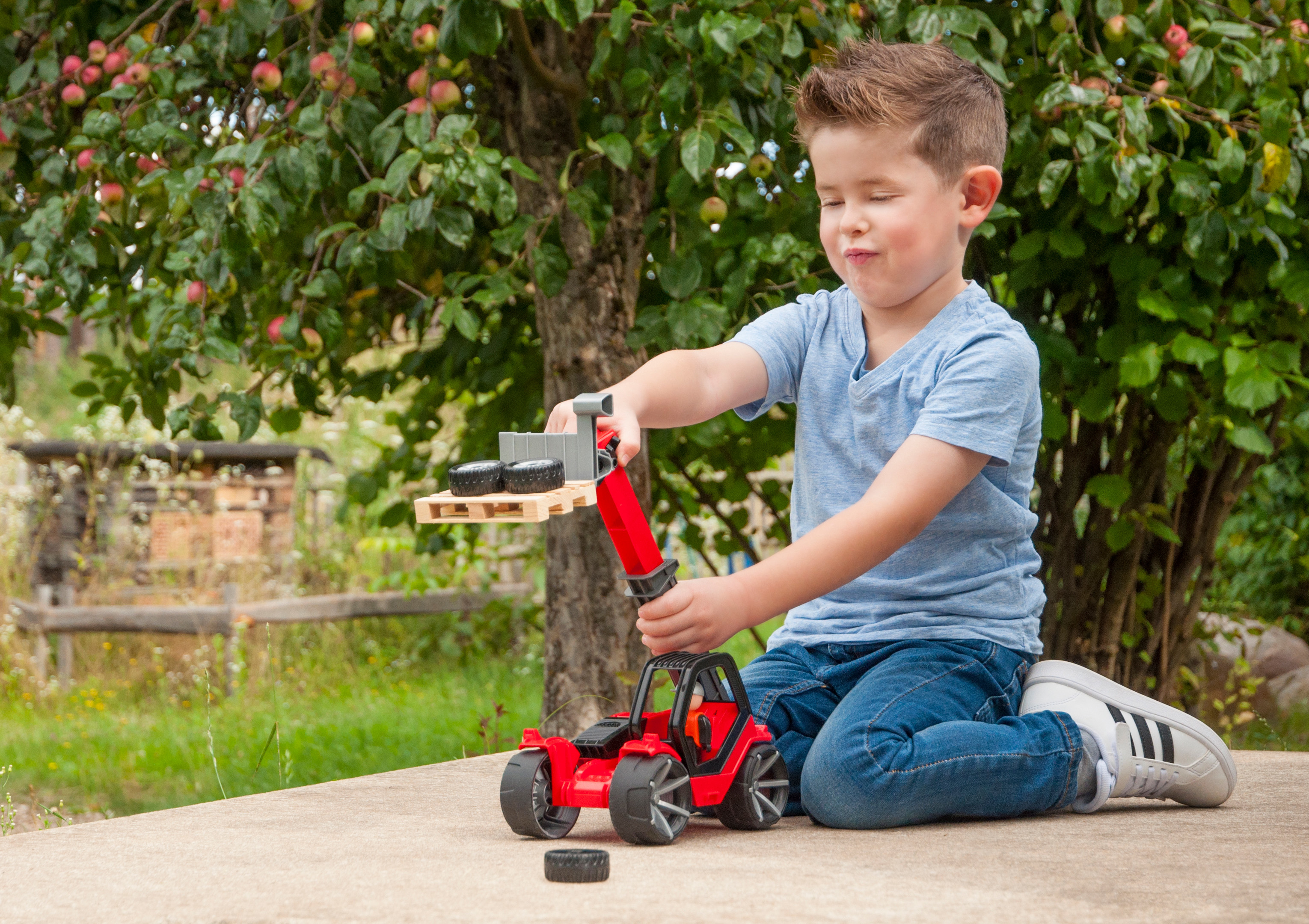Ein kleiner Junge spielt mit einem Spielzeug-Gabelstapler und bewegt Reifen. Er sitzt auf Beton.