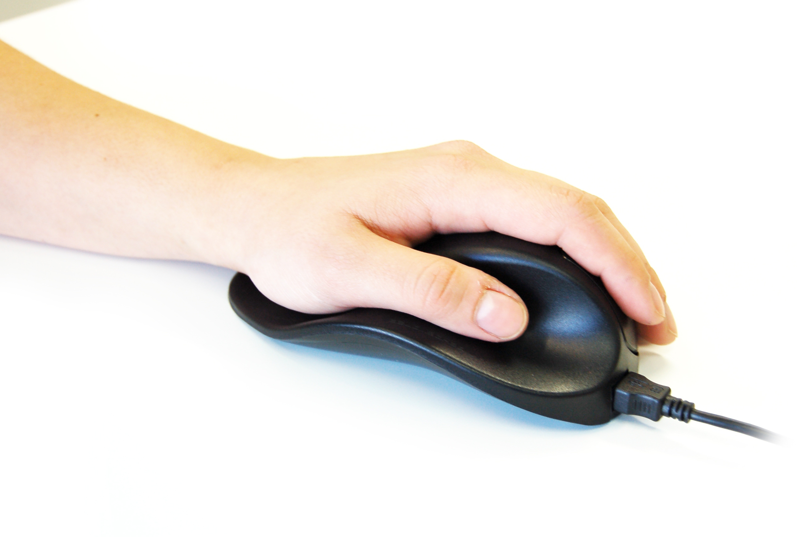 La mano de una persona sosteniendo un ratón ergonómico negro sobre una superficie blanca.