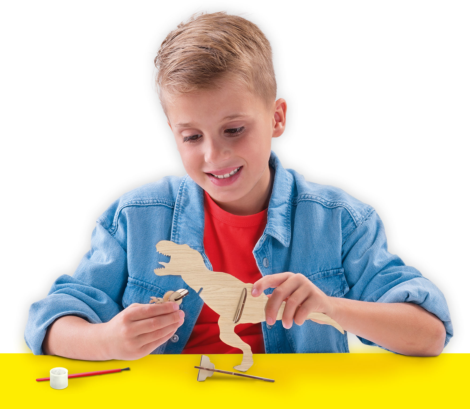 Chłopiec składający drewnianą zabawkę dinozaura. Żółty stół, białe tło.
