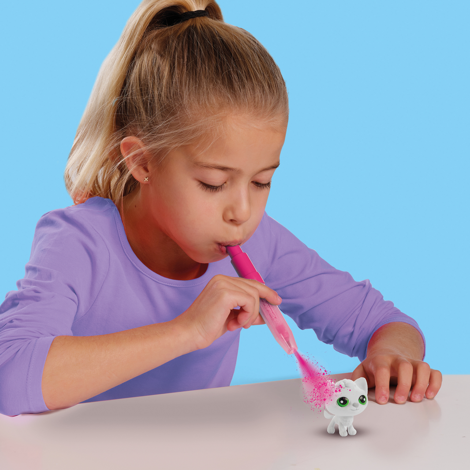 Dziewczyna dmucha różowym proszkiem na białą figurkę kota. Niebieskie tło.