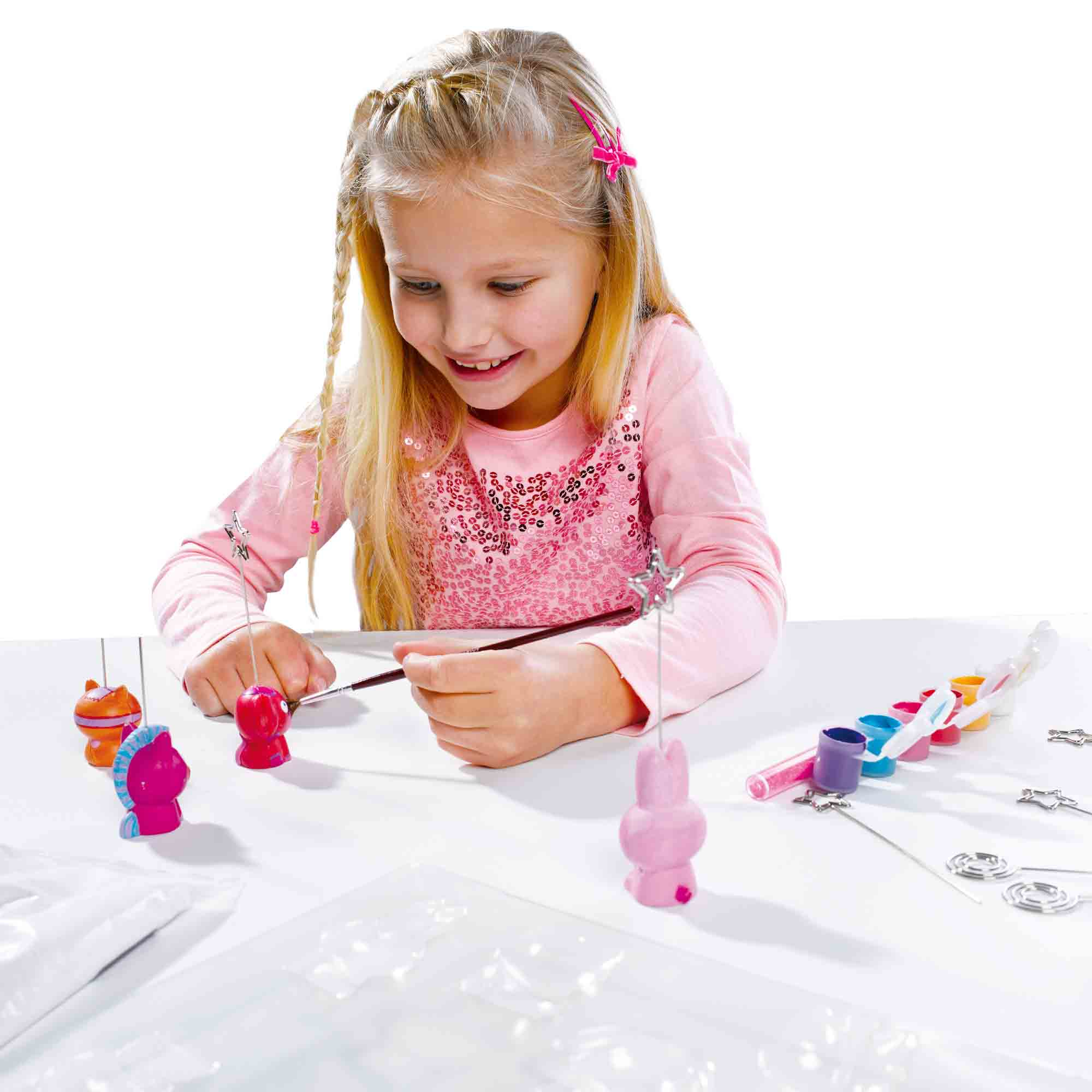 Dziewczyna maluje małe plastikowe figurki na białym stole pędzlem. Widocznych jest kilka figurek i pojemników z farbą.