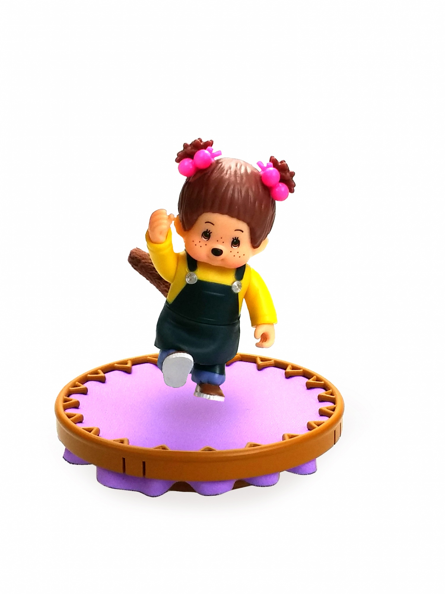 Figurka małpy z warkoczykami, stojąca na fioletowej platformie, z brązową podstawą. Białe tło.