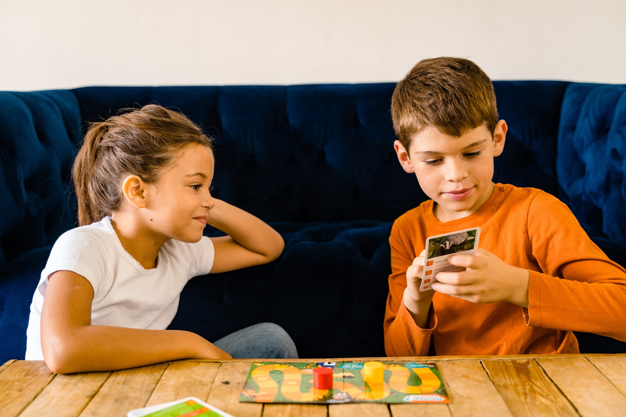 Dwoje dzieci grających w grę planszową na stole. Niebieska sofa w tle.