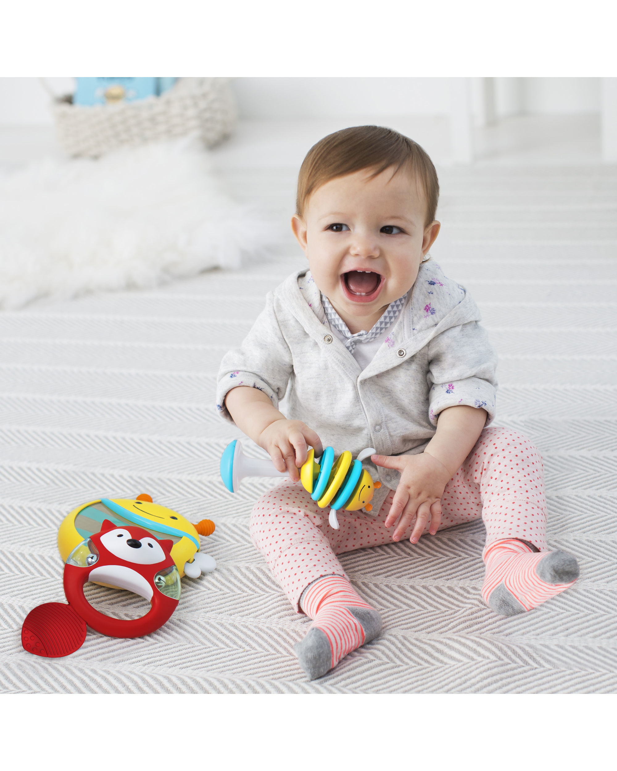 Ein Baby sitzt auf einem Teppich und spielt mit Spielzeug. Spielzeug ist ein Bienen-Shaker, ein Fuchspielzeug und eine Rassel.