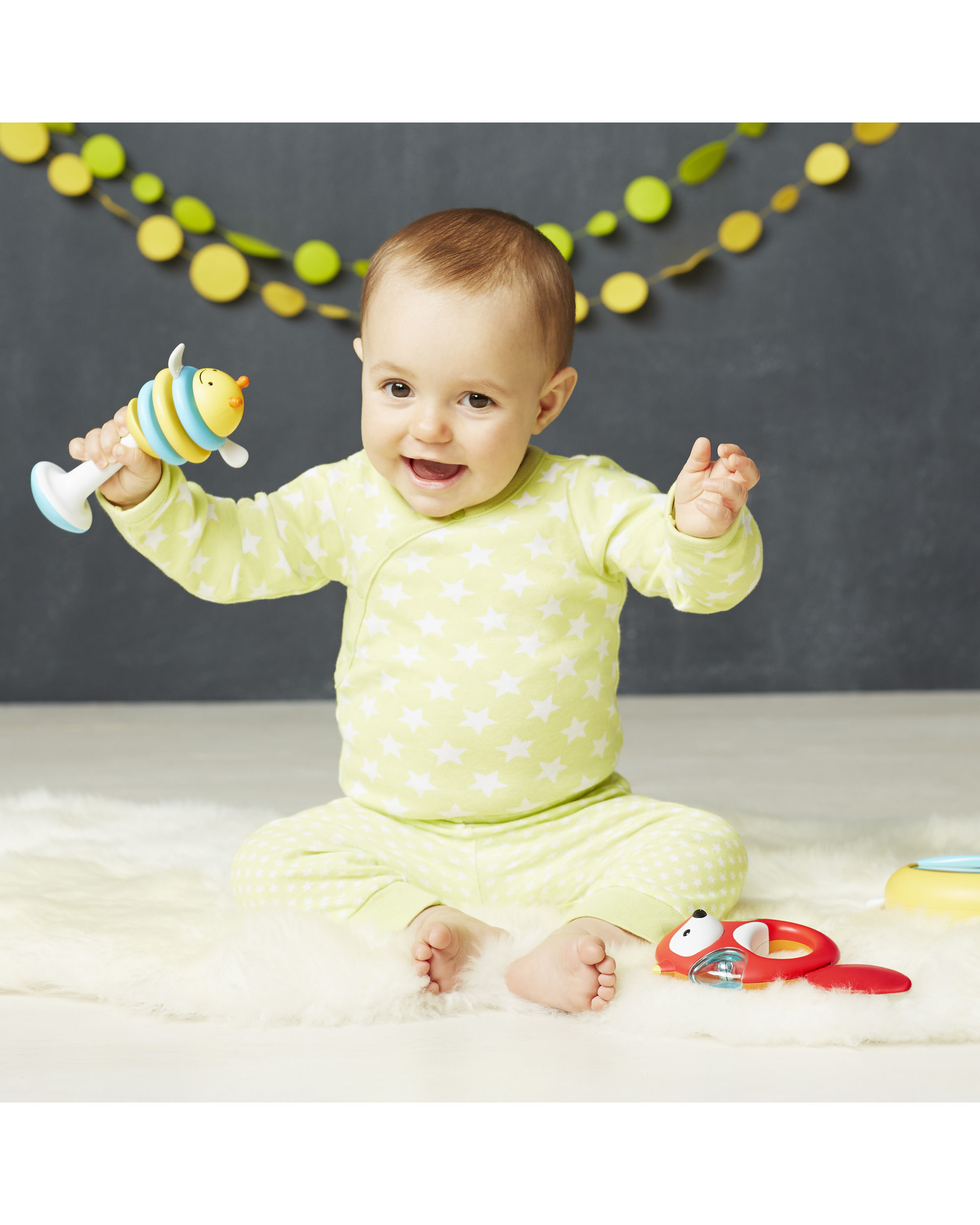 Ein Baby sitzt und hält ein buntes Spielzeug. Es gibt einen Teppich und andere Spielsachen im Hintergrund.