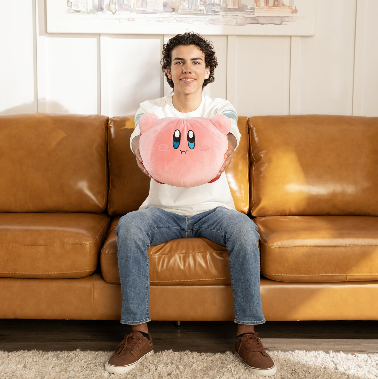 Młody mężczyzna trzymający różową pluszową zabawkę Kirby, siedzący na brązowej skórzanej sofie w pokoju.