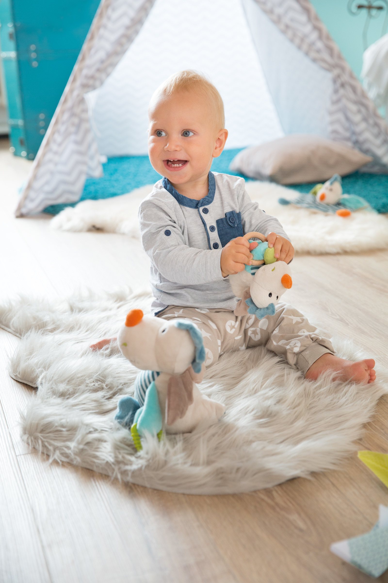 Glückliches Baby sitzt auf einem Teppich. Spielzeug und Zelt im Hintergrund. Lächelt und greift nach einem Spielzeug.