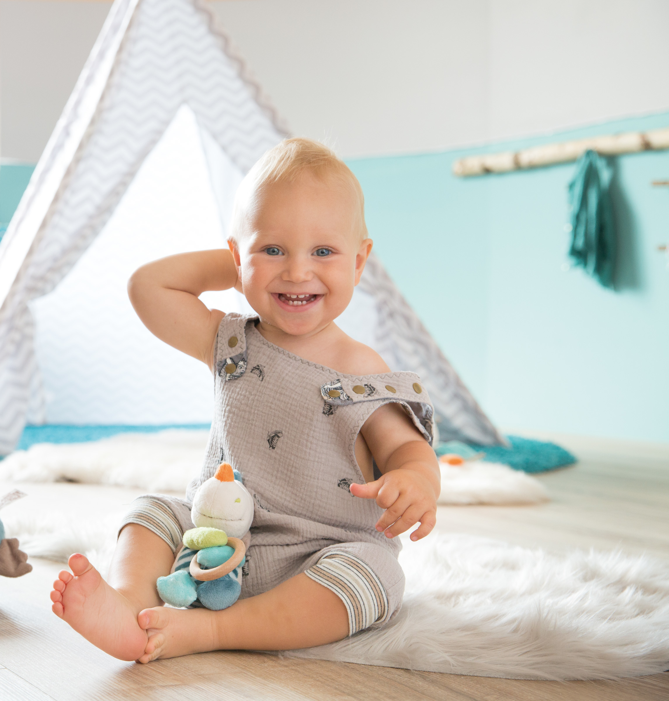 Baby sitzt auf einem Teppich. Lächelt und spielt mit einem Spielzeug. Zelt im Hintergrund.