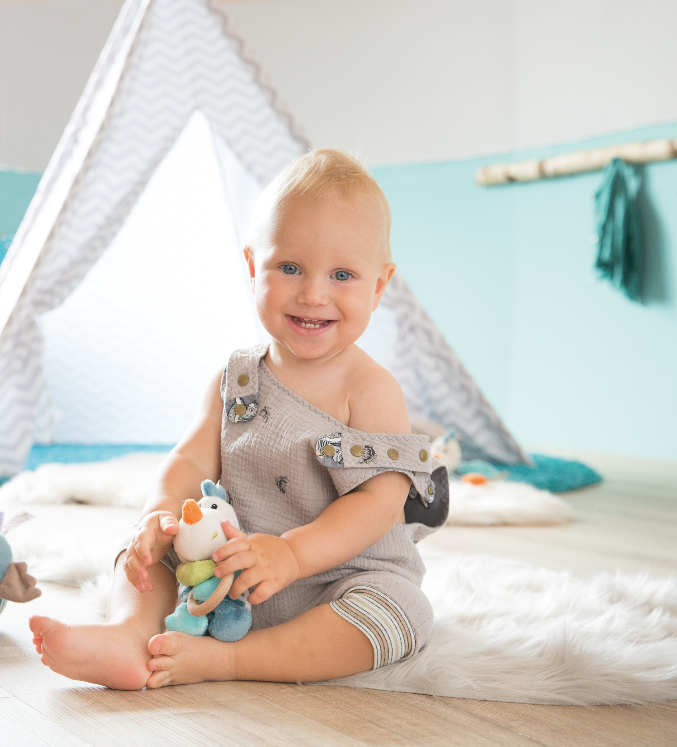 Ein lächelndes Baby sitzt auf einem flauschigen Teppich und hält ein Spielzeug. Ein Zelt ist im Hintergrund.