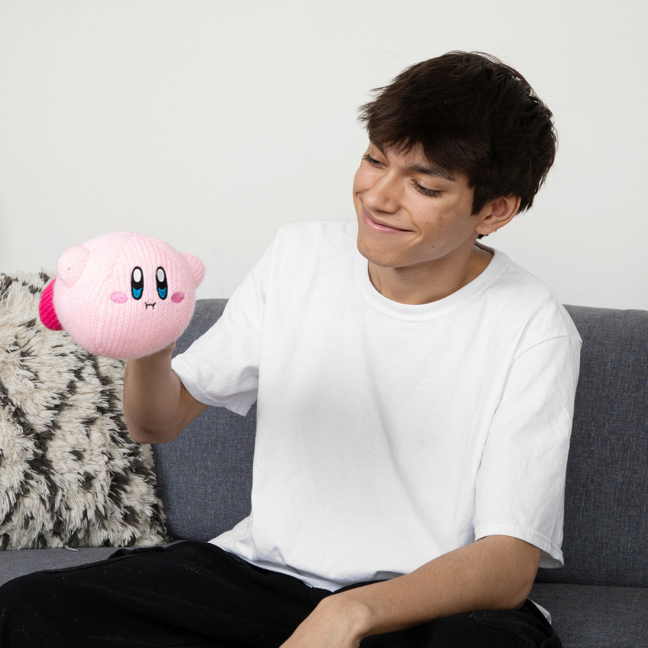 Un giovane sorride, tenendo un peluche rosa di Kirby. È seduto su un divano grigio contro un muro bianco.
