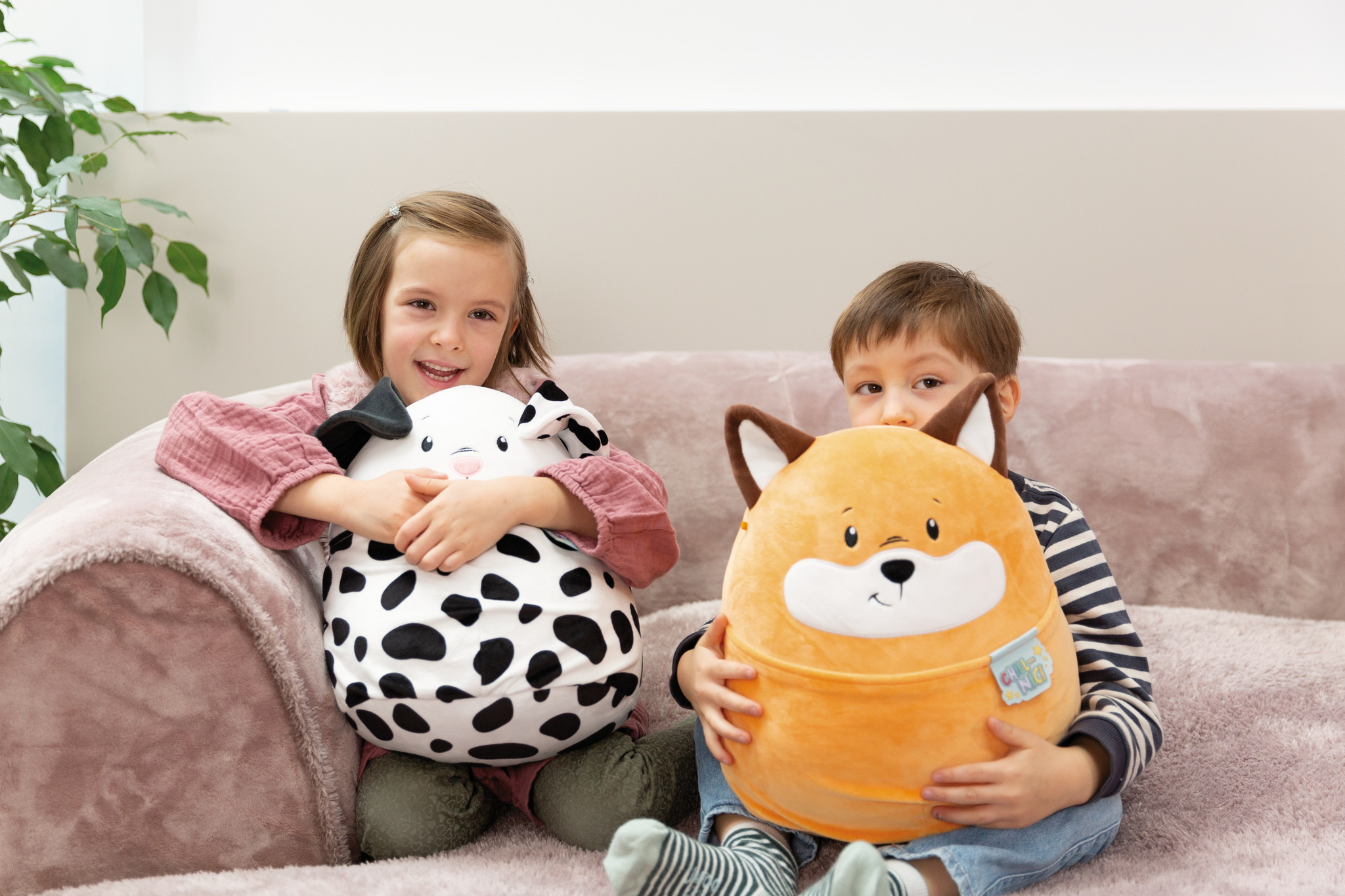 Zwei Kinder, eines hält ein Kuh-Plüschtier, das andere ein Fuchs-Plüschtier, lächeln auf einem rosa Sofa. Helles Umgebungslicht.