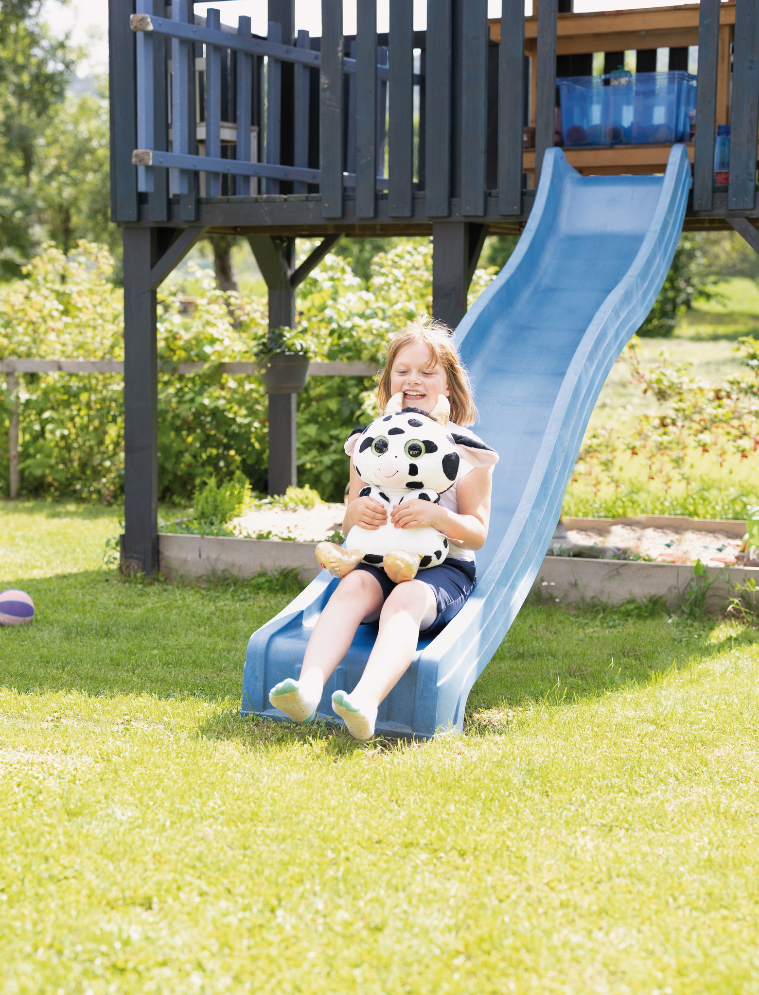 Ein Mädchen auf einer blauen Rutsche, das ein Plüschkuhspielzeug hält. Die Szene spielt in einem grasbewachsenen Garten mit einer Holzkonstruktion im Hintergrund.