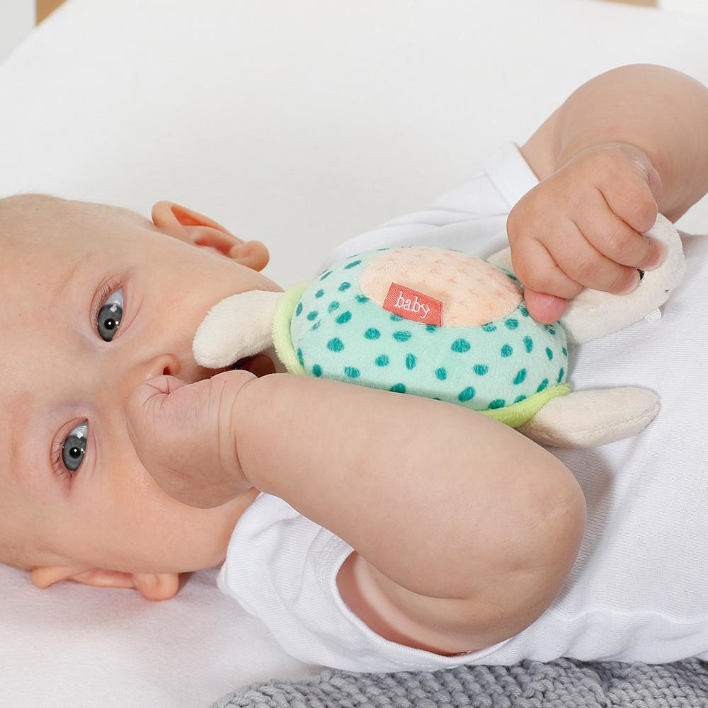 Niemowlę leżące na białej powierzchni, trzymające zabawkę w kształcie żółwia. Dziecko patrzy na zabawkę.