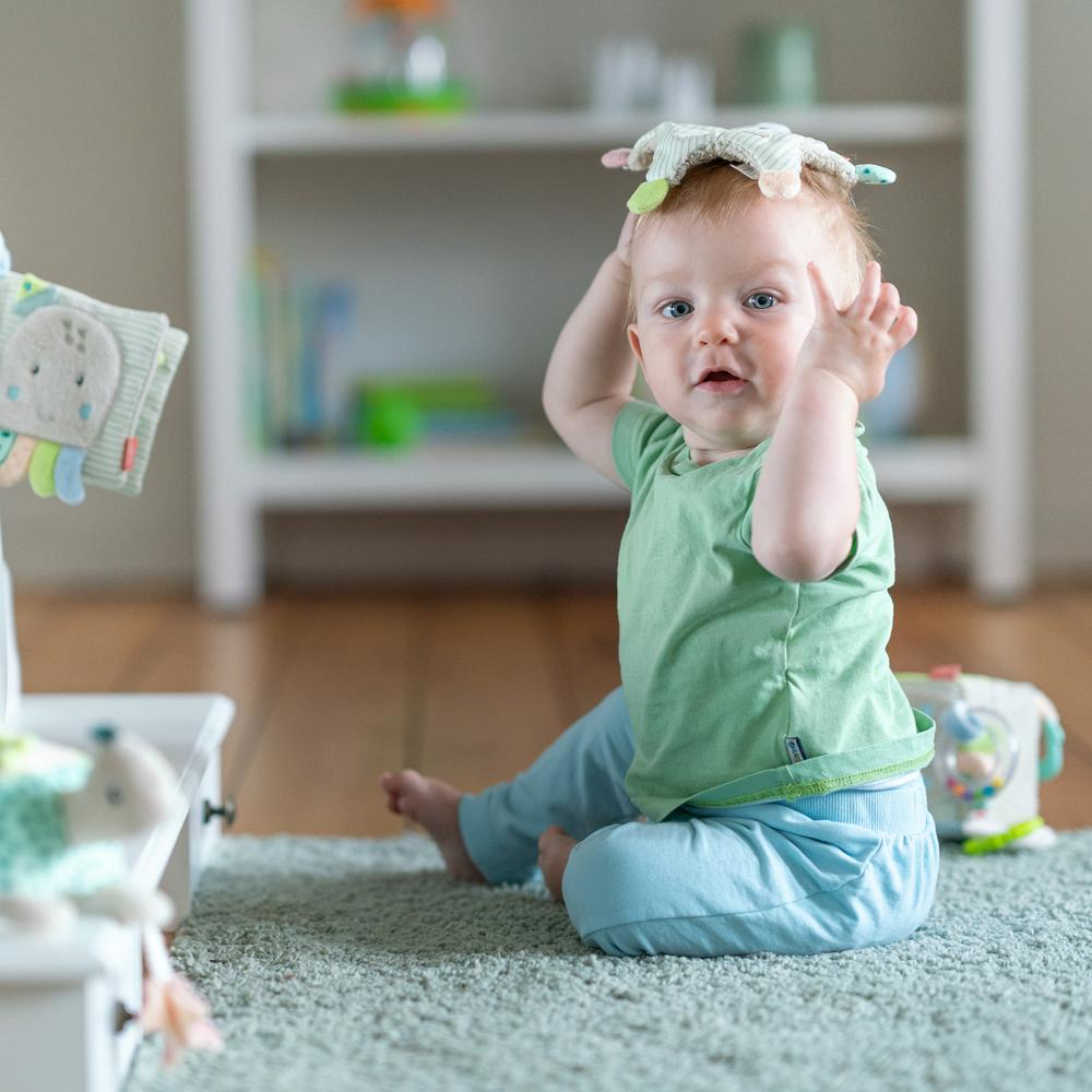 Niemowlę siedzi na dywanie i bawi się zabawkami. Widoczna jest karuzela i książeczka dla dzieci.