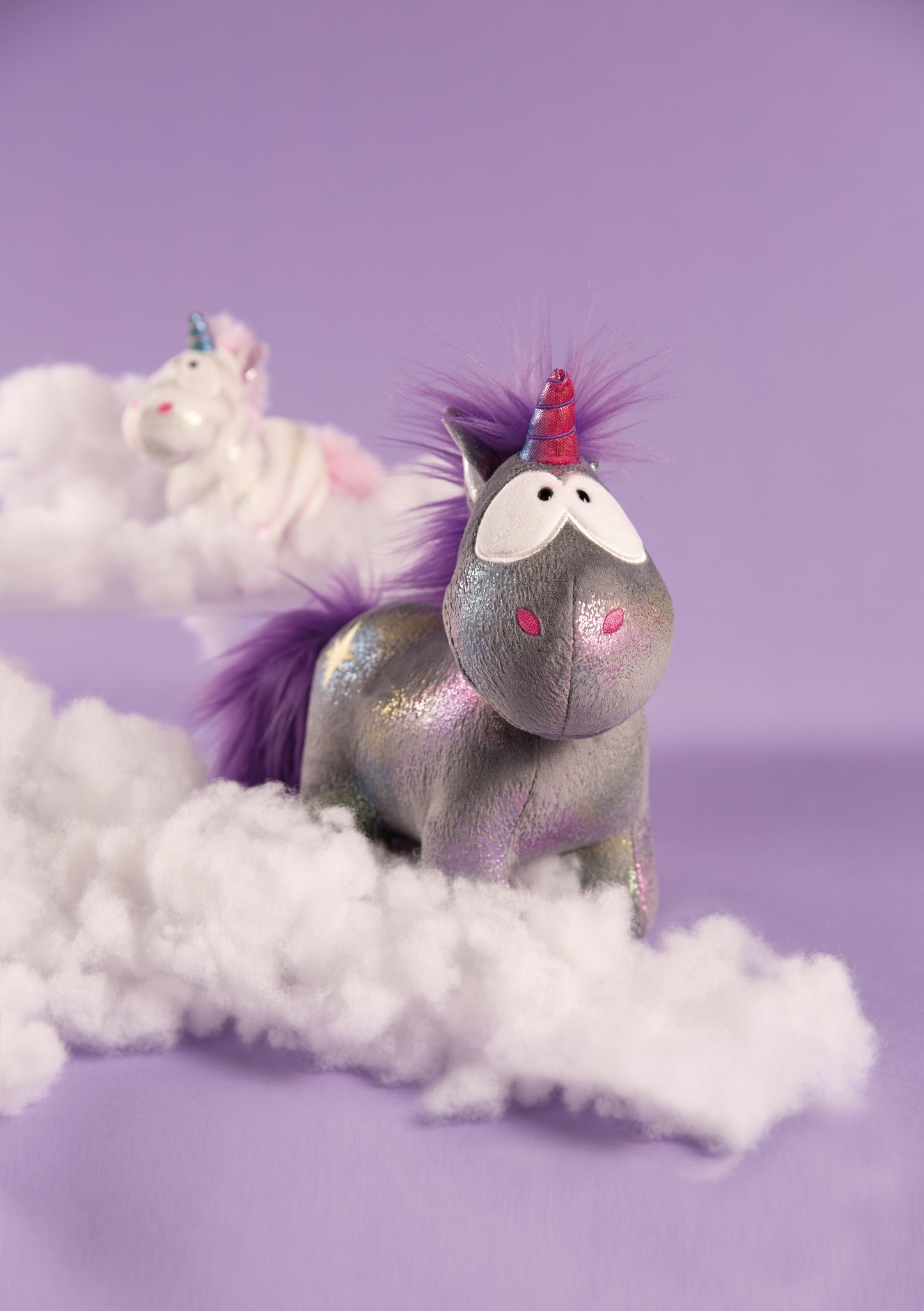 Dwie pluszowe zabawki jednorożce na chmurach, fioletowe tło. Jeden jest blisko, drugi w tle.