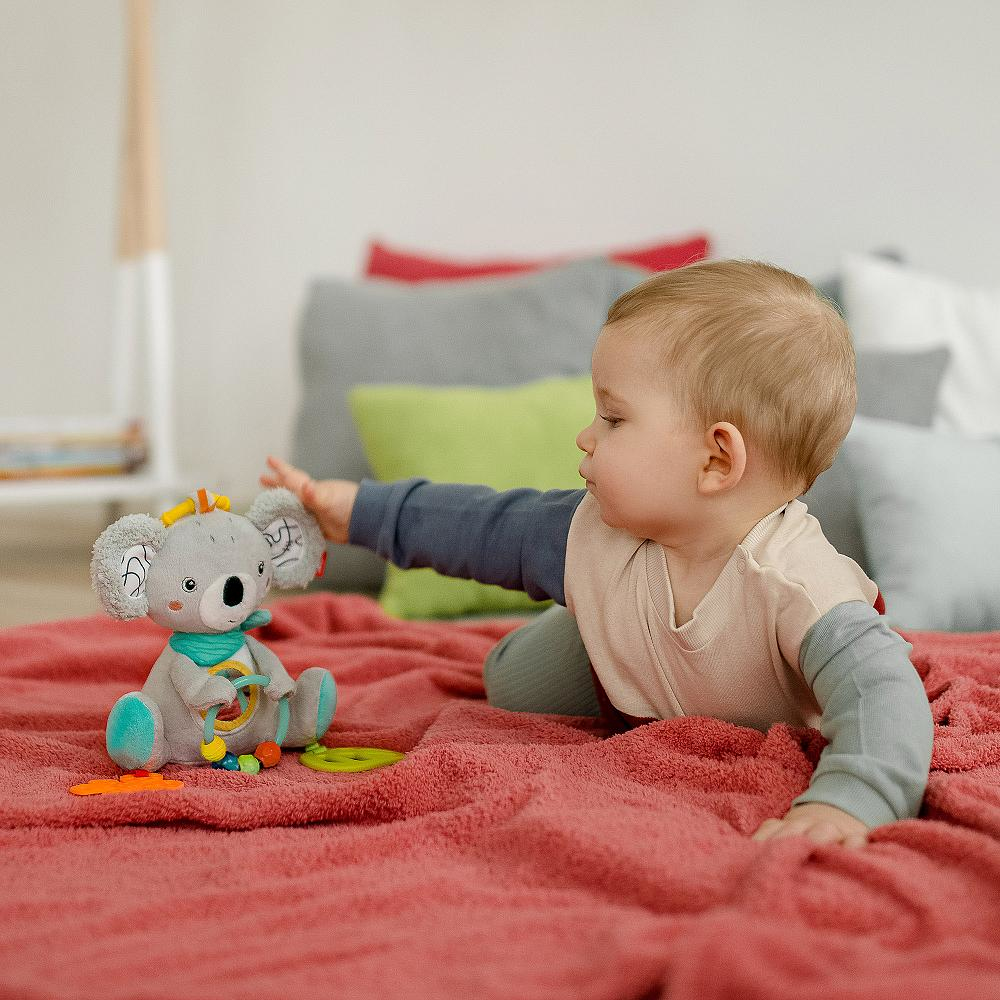 Baby greift nach einem Koala-Spielzeug auf einer roten Decke. Hintergrund: Kissen und eine Wand.