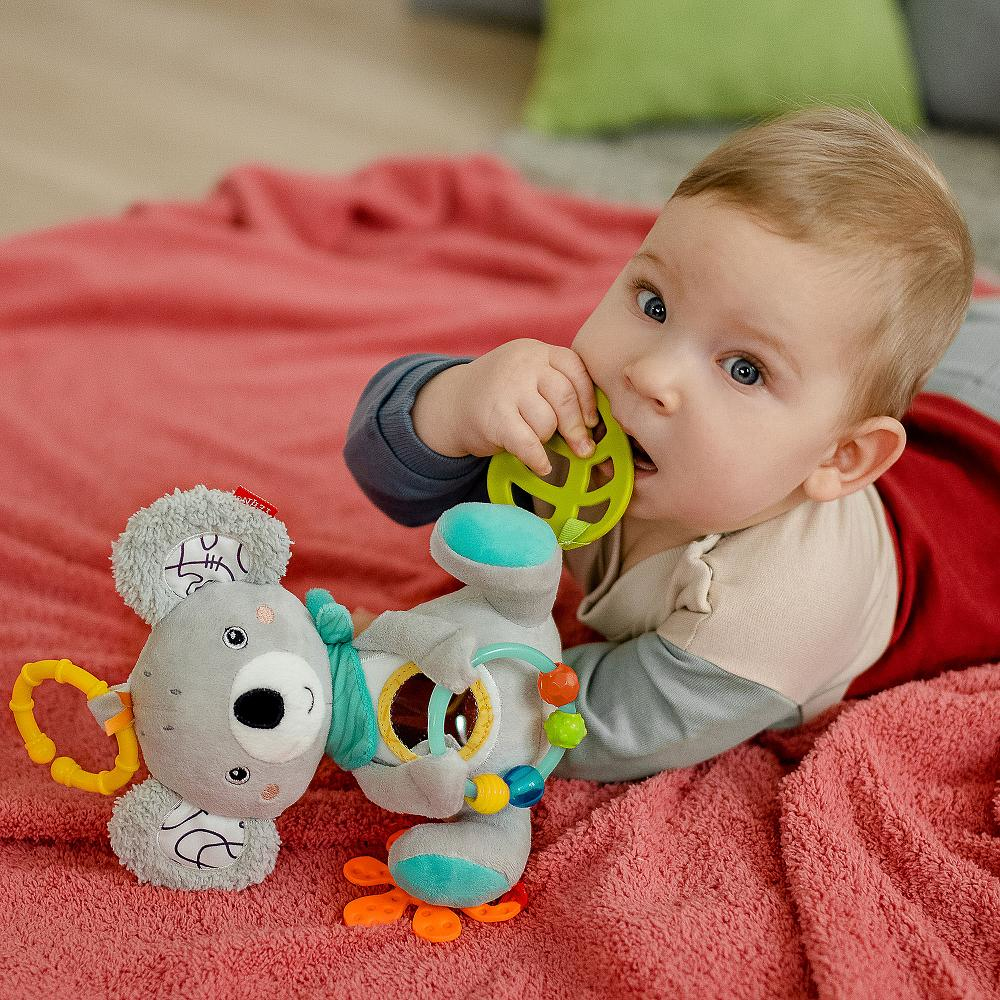 Baby liegt auf einer rosa Decke, mit einem Spielzeug im Mund und einem Stoff-Koala-Spielzeug daneben.