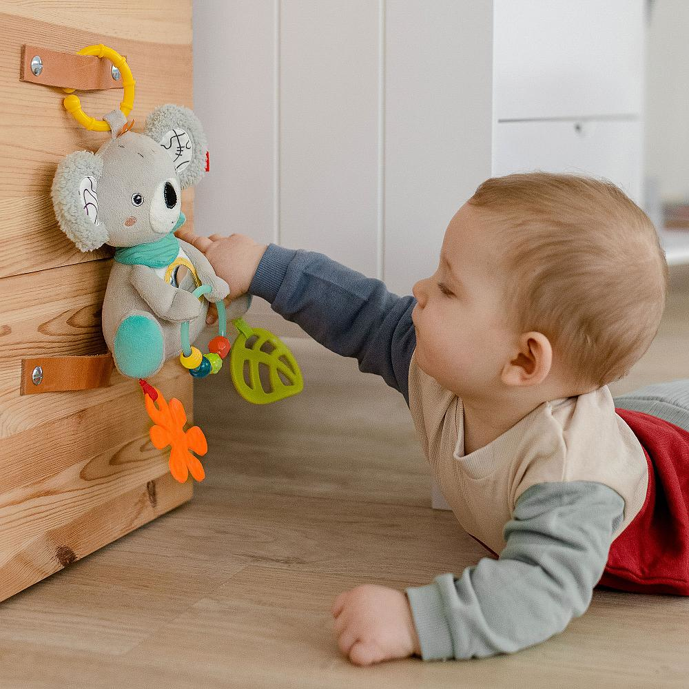 Baby greift nach Koala-Spielzeug an Holzkonstruktion. Hintergrund: Holzwand und -boden.