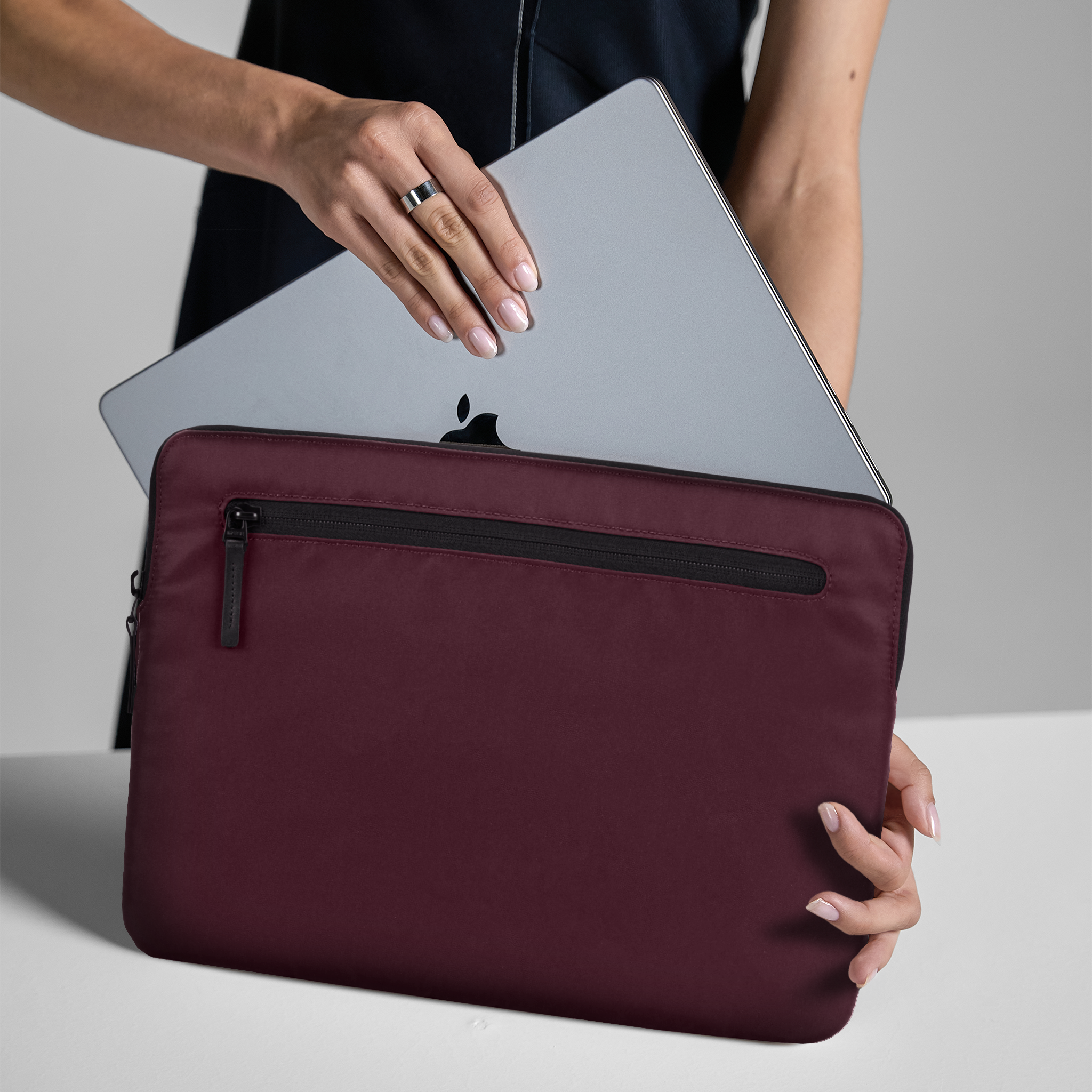 Una persona che tiene un laptop e una custodia bordeaux su una superficie bianca, con sfondo neutro.