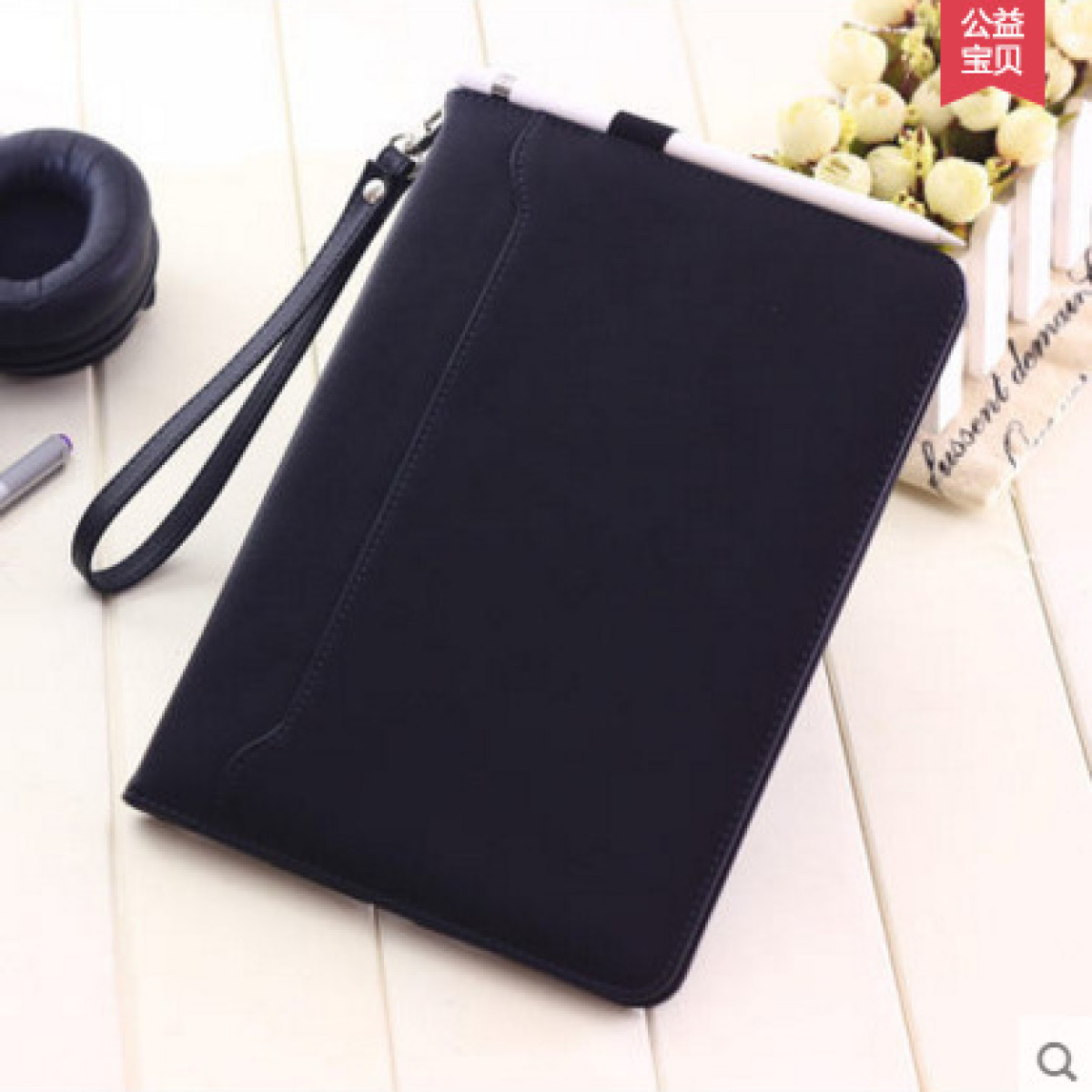 Custodia nera per tablet con cinturino da polso. Una penna e cuffie sullo sfondo.