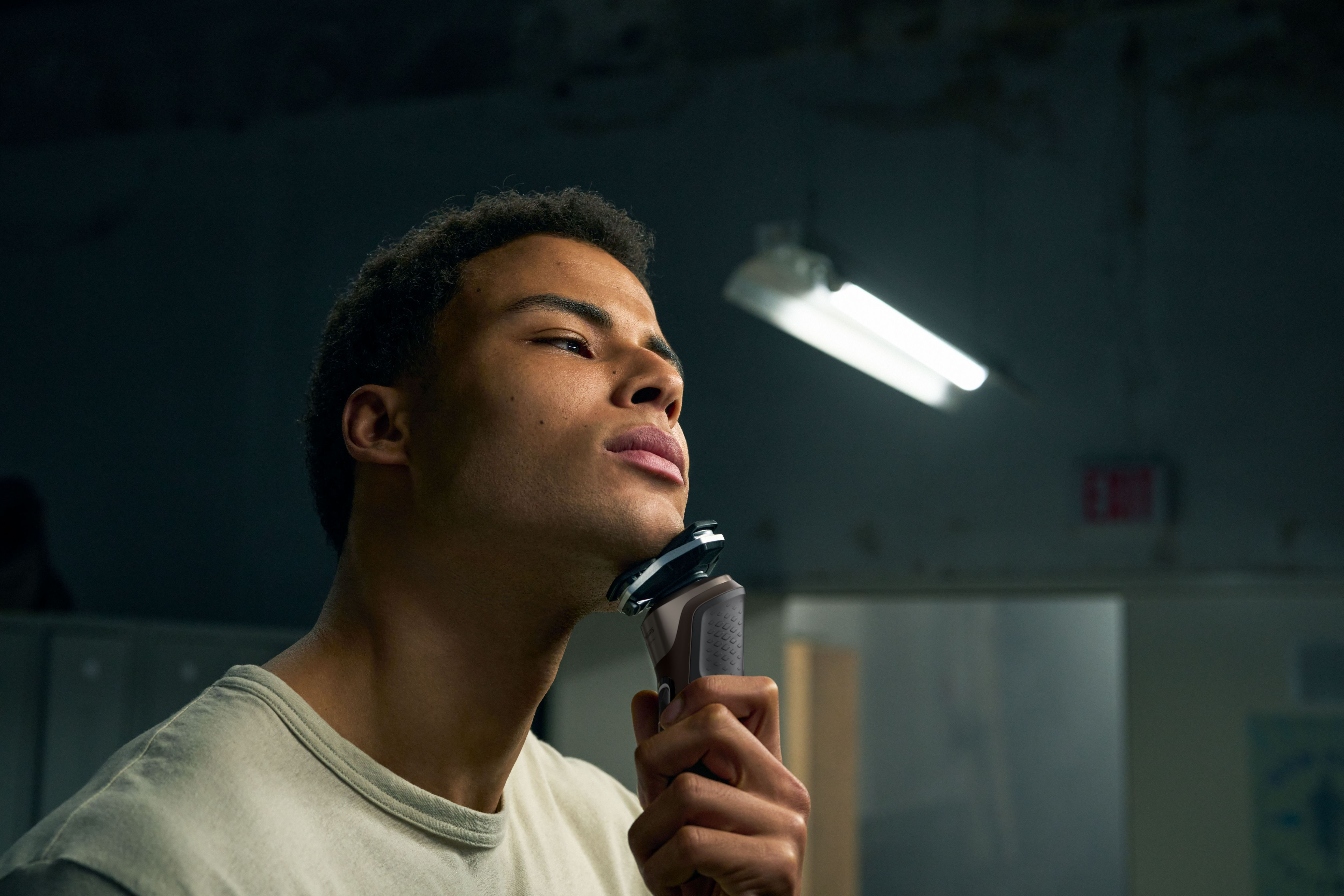 Mężczyzna goli policzek golarką Philips. Słabe oświetlenie, tło przedstawia ścianę i oprawę oświetleniową.