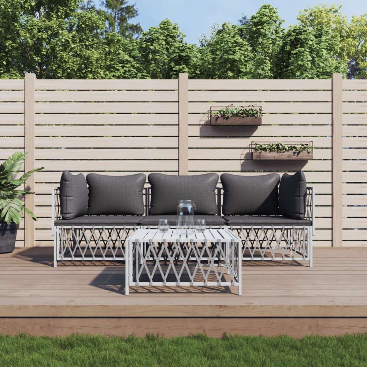 Terrassenset mit grauen Kissen, weißem Rahmen und passendem Tisch auf Holzterrasse. Grüne Pflanzen und Zaun.