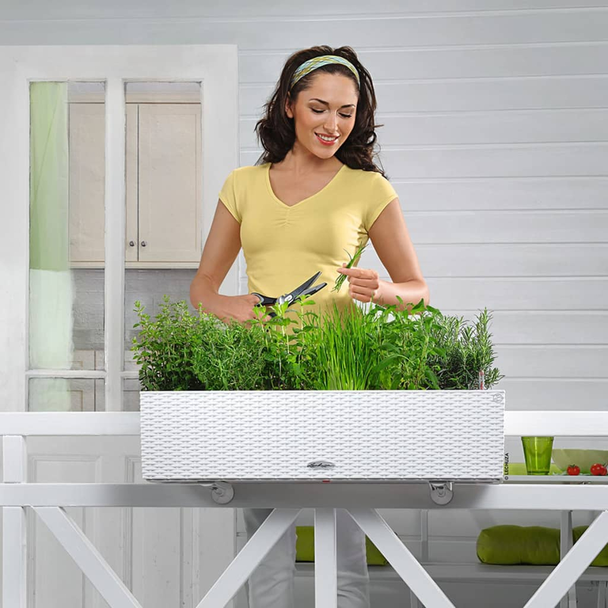 Une femme en jaune coupe des herbes dans un bac à plantes blanc sur un balcon. Des plantes vertes.