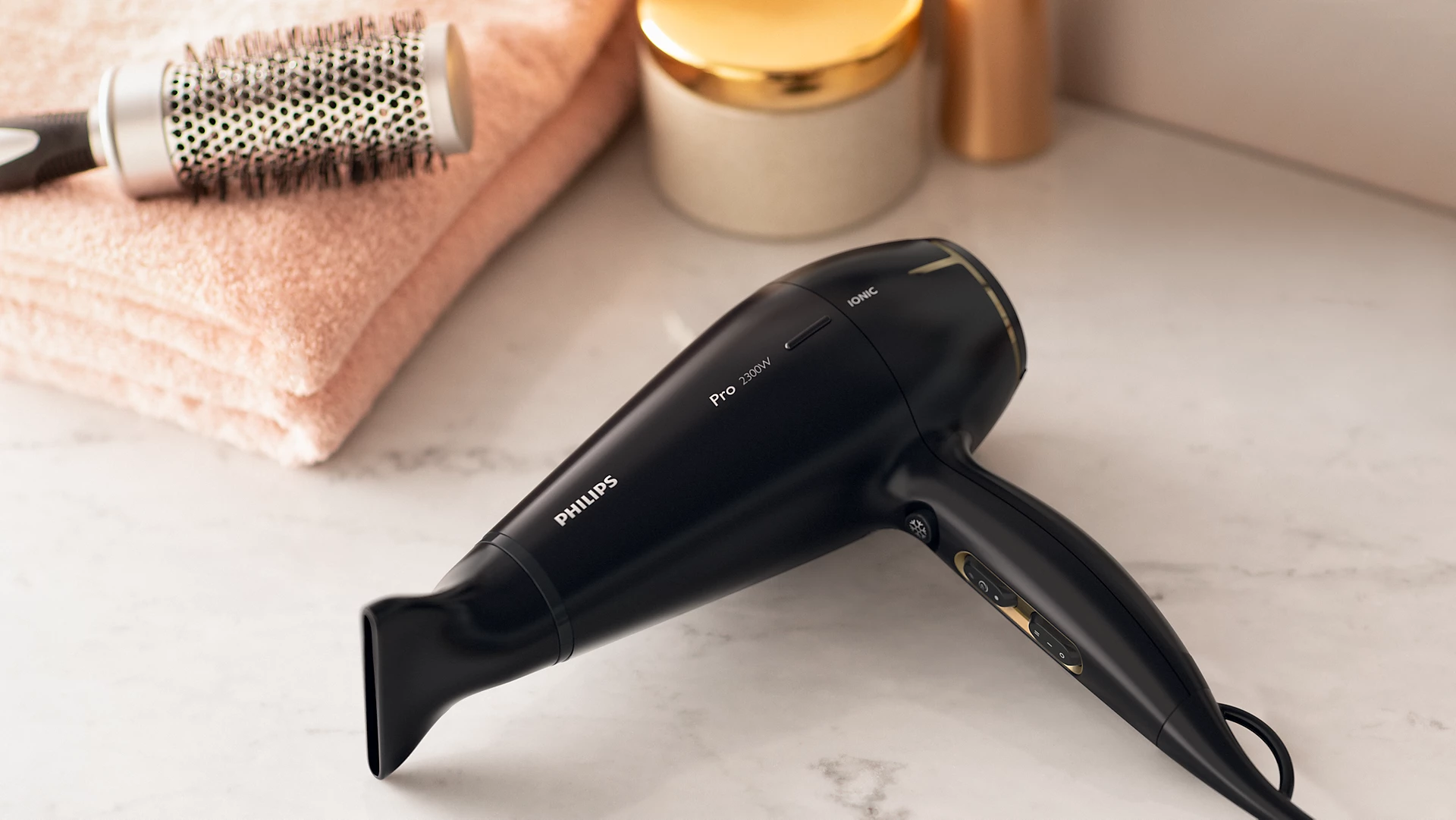 Czarna suszarka do włosów Philips na marmurowej powierzchni, z szczotką i ręcznikiem w tle. Widoczne są również słoiki.