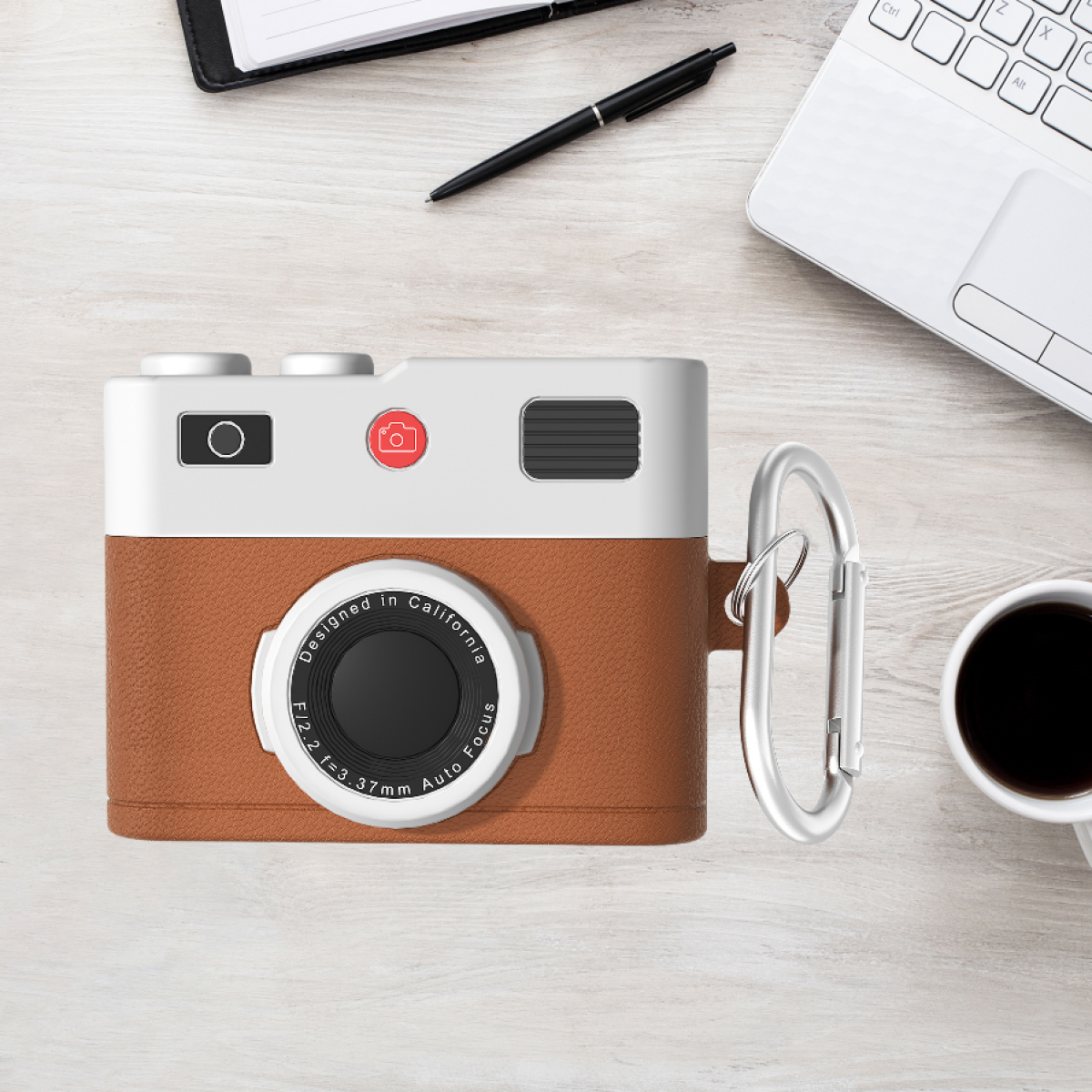 Una custodia per AirPod a forma di fotocamera su una scrivania con un laptop e una tazza di caffè.