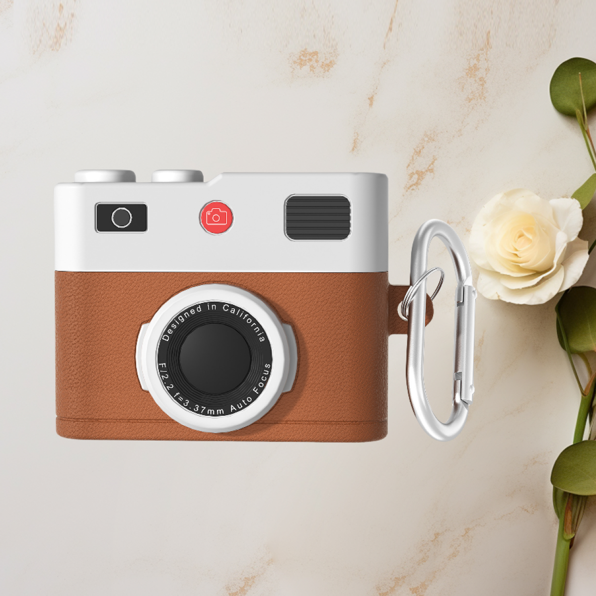 Custodia a forma di fotocamera con parte anteriore marrone, parte superiore bianca e moschettone argentato. Rosa e foglie vicine.