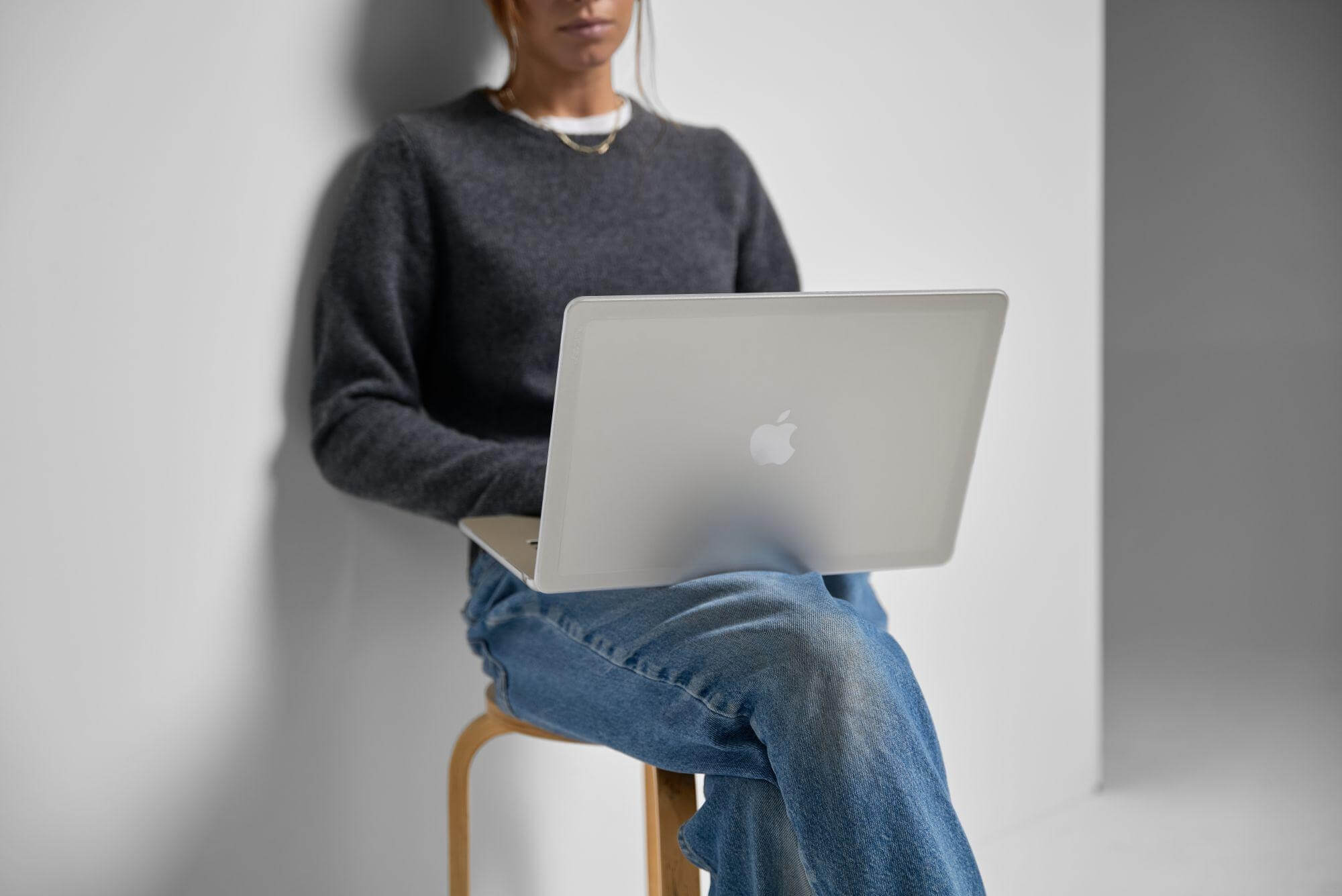 Una persona seduta con un laptop argentato sulle ginocchia. Indossa un maglione grigio e jeans.