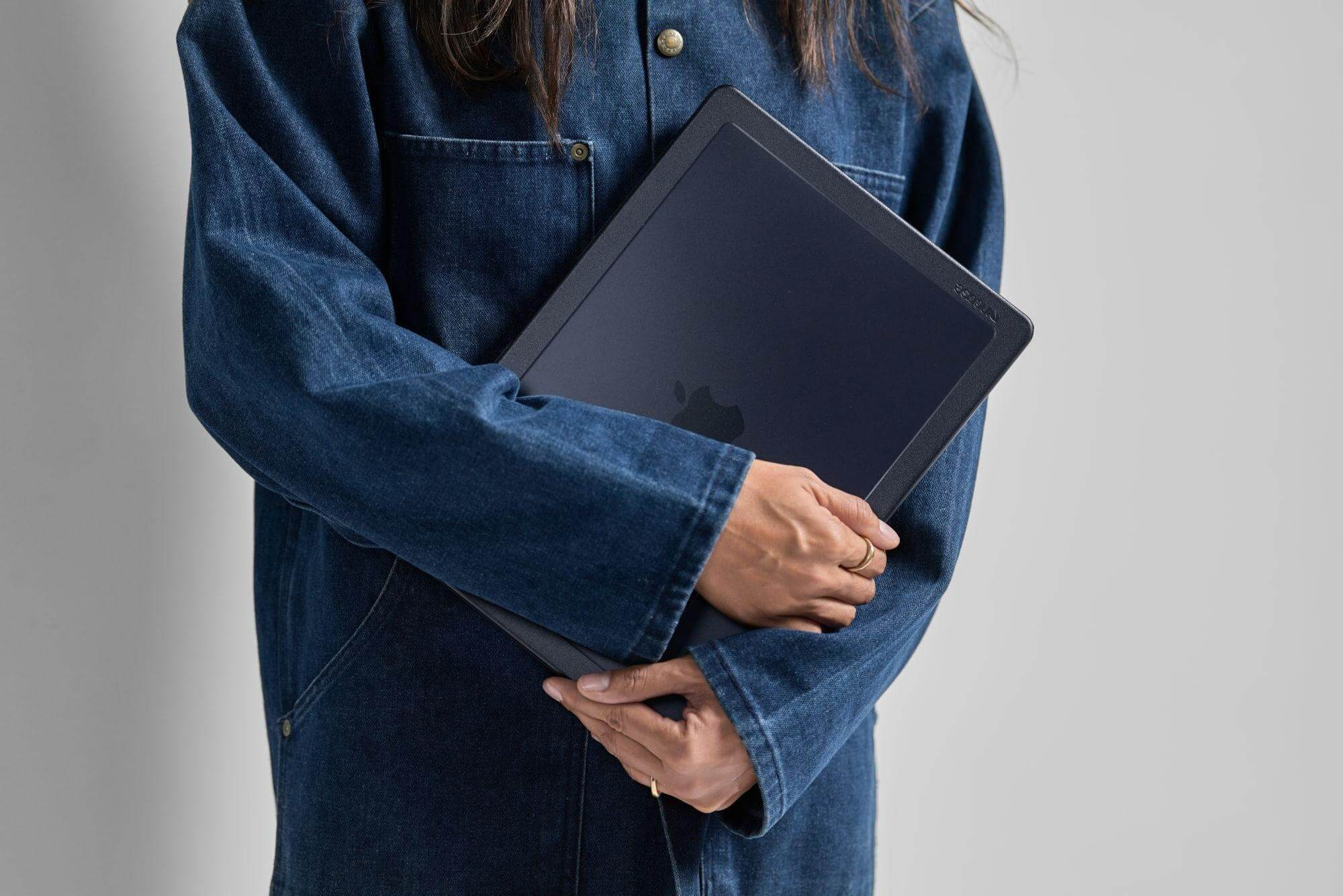 Eine Person in einer Jeansjacke hält einen geschlossenen dunklen Laptop. Das Apple-Logo ist auf dem Deckel sichtbar.