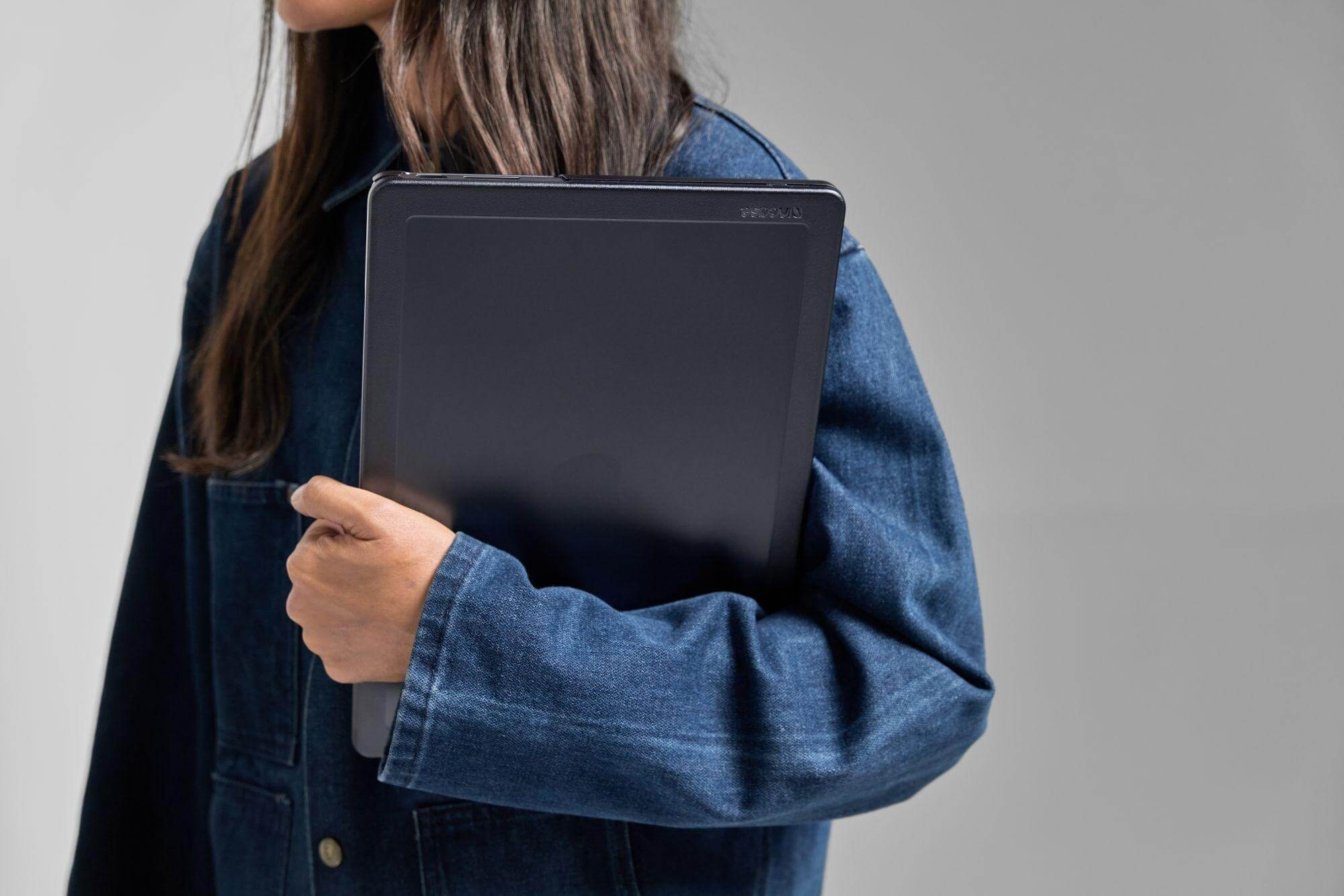 Une femme tenant un ordinateur portable dans un étui gris foncé, portant une veste en jean.