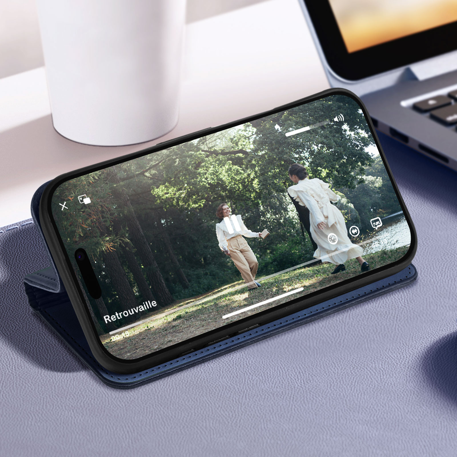 Telefono in custodia blu, riproduce un video. Tazza bianca e laptop sullo sfondo. Superficie blu.