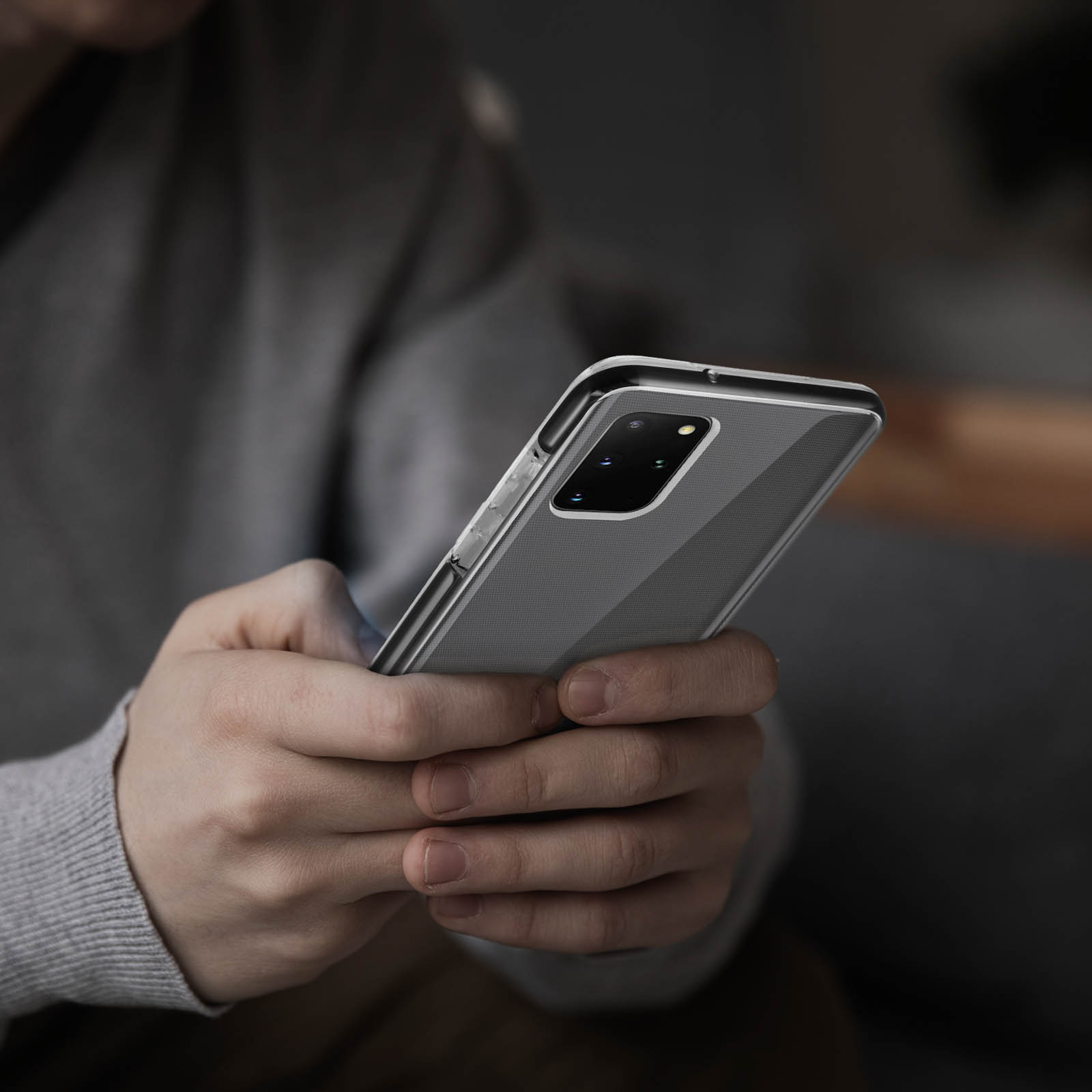 Mano che tiene un telefono con custodia trasparente. Il telefono ha tre fotocamere. Sfondo scuro.