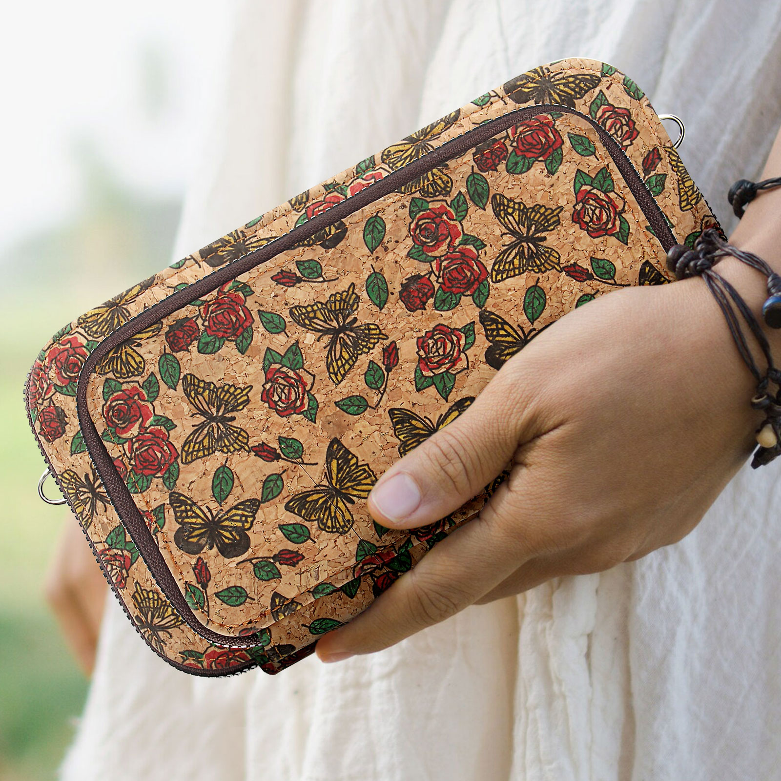 Person hält eine Korkbrieftasche mit Schmetterlings- und Rosenmuster. Reißverschluss, braune Verkleidung. Armband sichtbar.