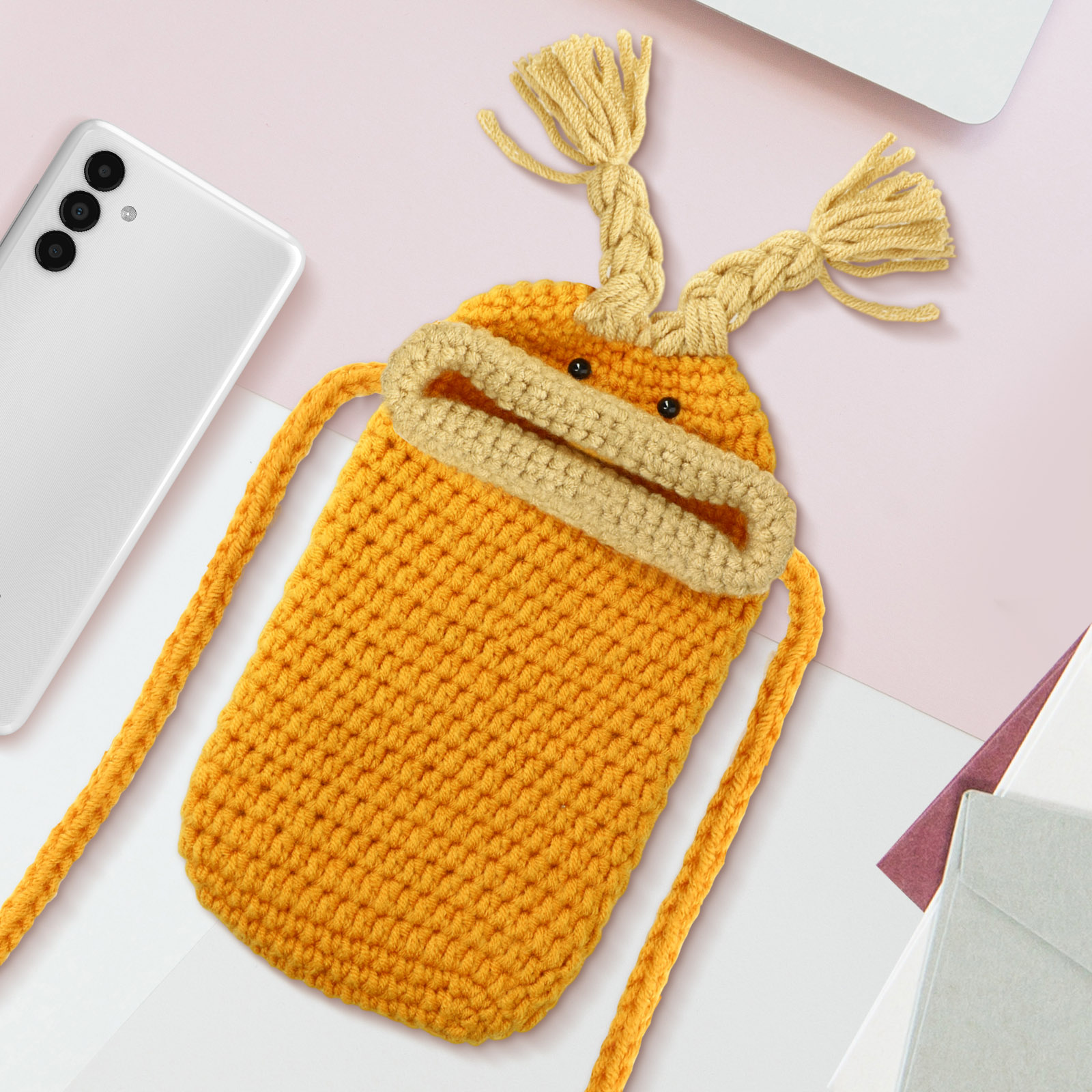 Borsa per telefono arancione lavorata a maglia con viso su scrivania con telefono, fogli e laptop.