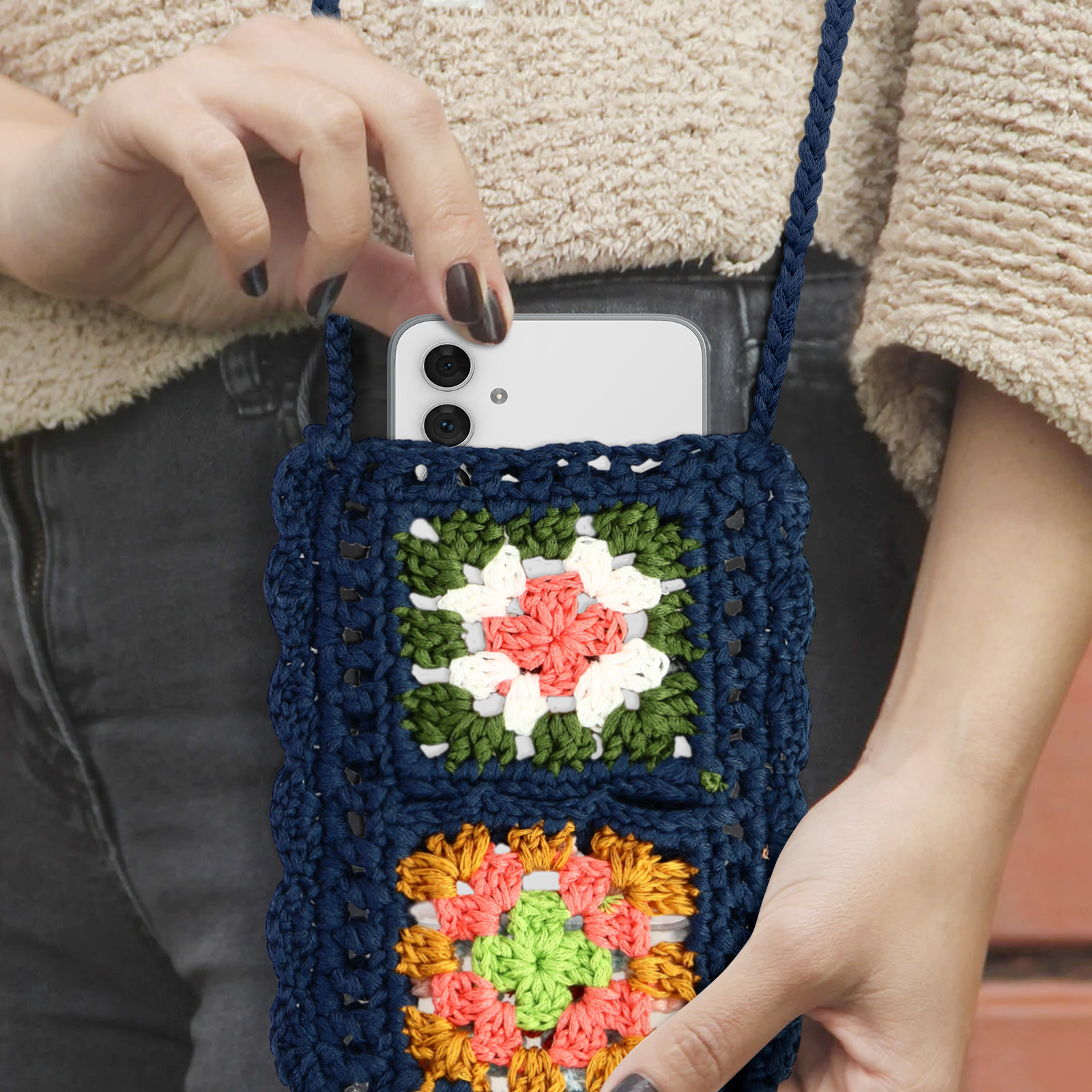 Le mani di una persona che tiene una borsa a tracolla all'uncinetto blu navy con un telefono dentro. La persona indossa una camicia beige.