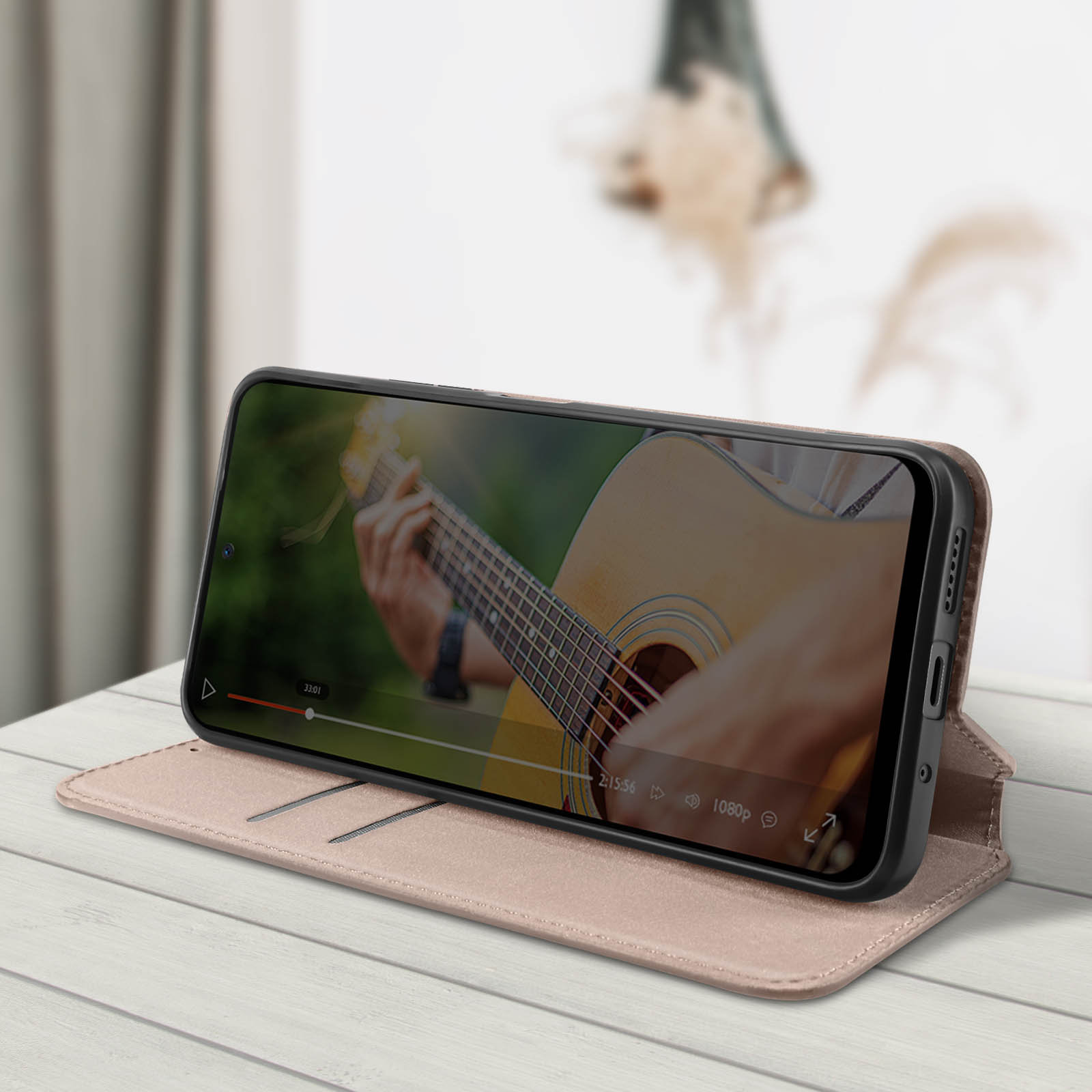 Telefono in custodia beige, video in riproduzione, scena con chitarra, su superficie in legno.
