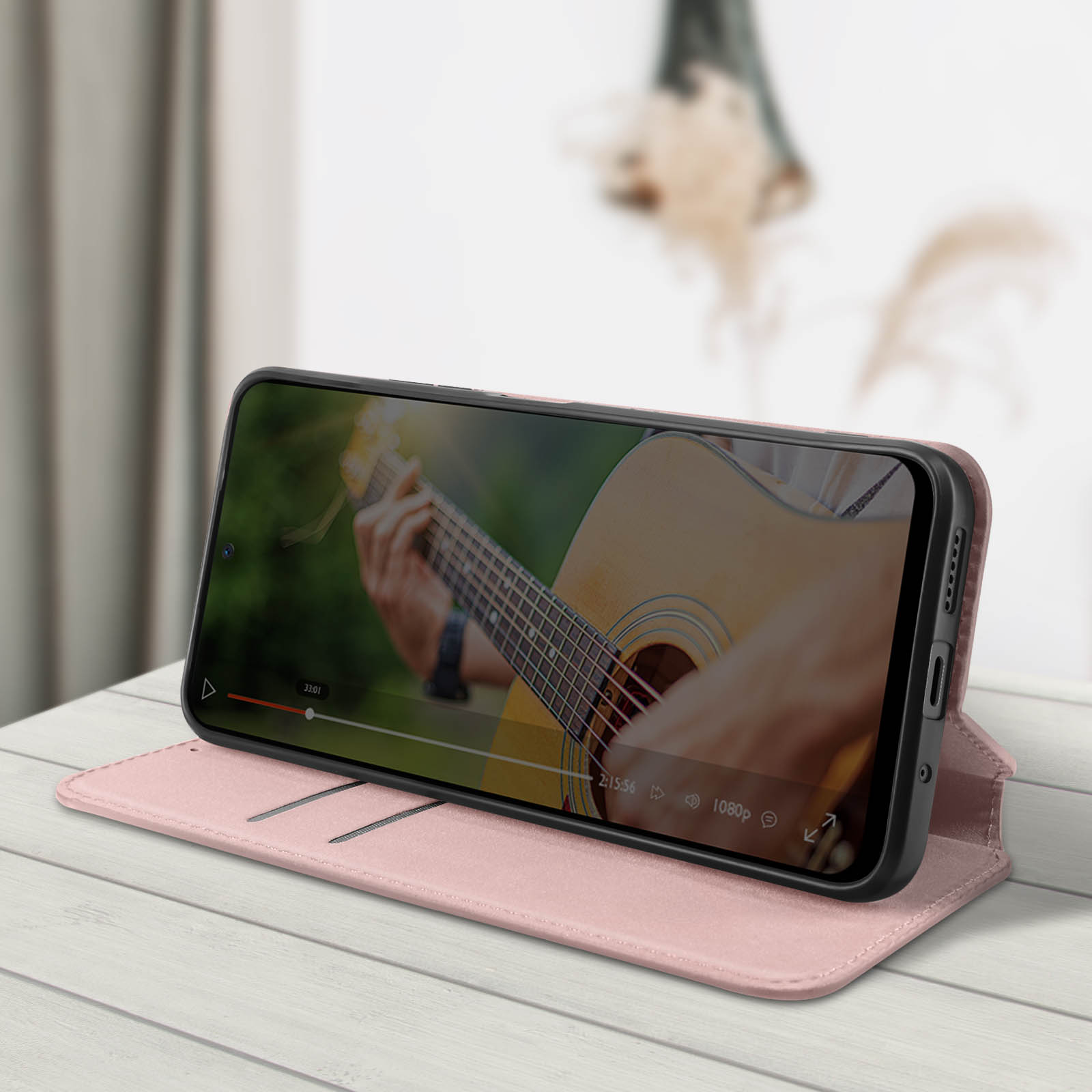 Teléfono en funda rosa, abierto, apoyado. Una persona toca la guitarra en la pantalla. Mesa de madera. Enfoque suave.
