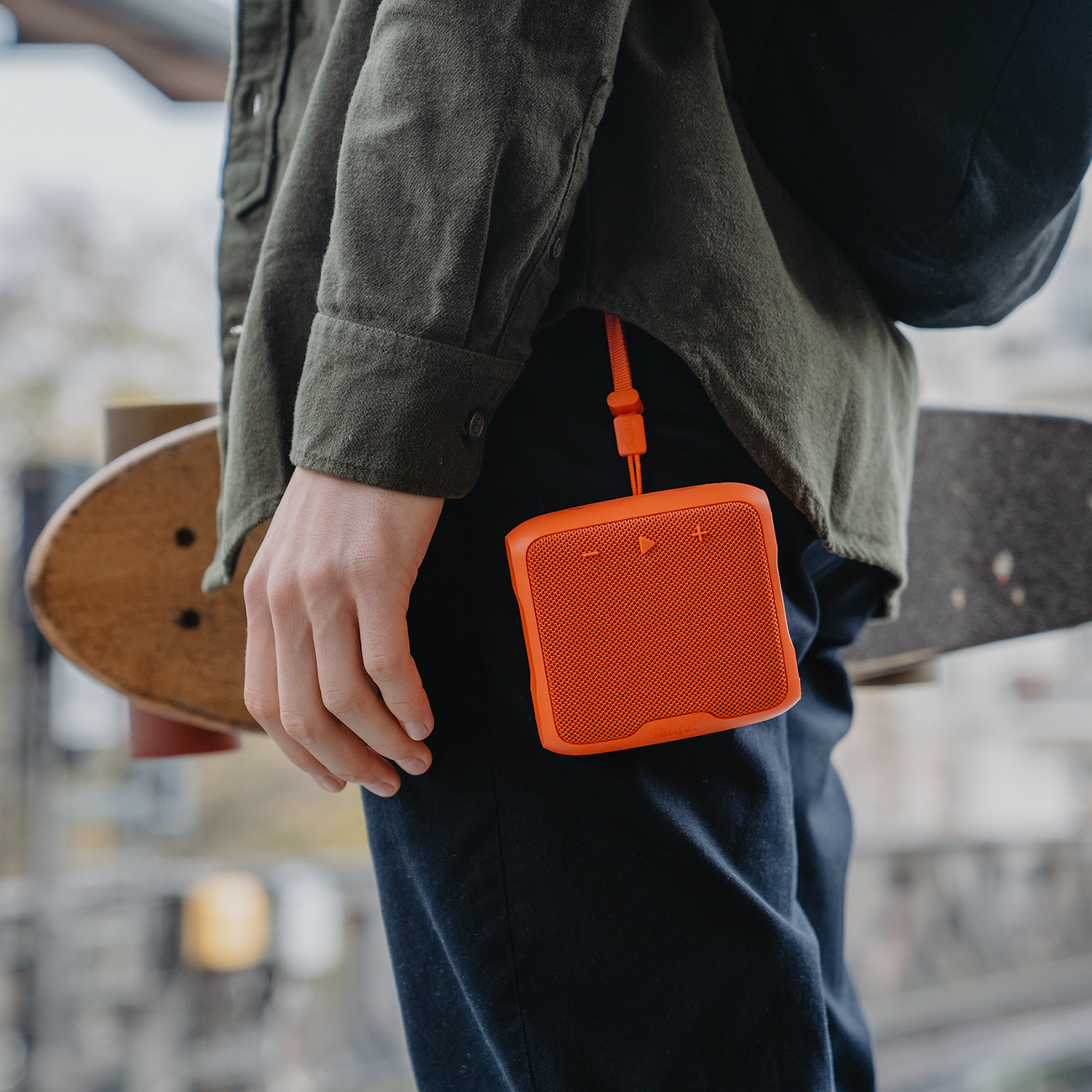 Persoon houdt oranje speaker vast, bevestigd aan broek. Skateboard en groen shirt zichtbaar.